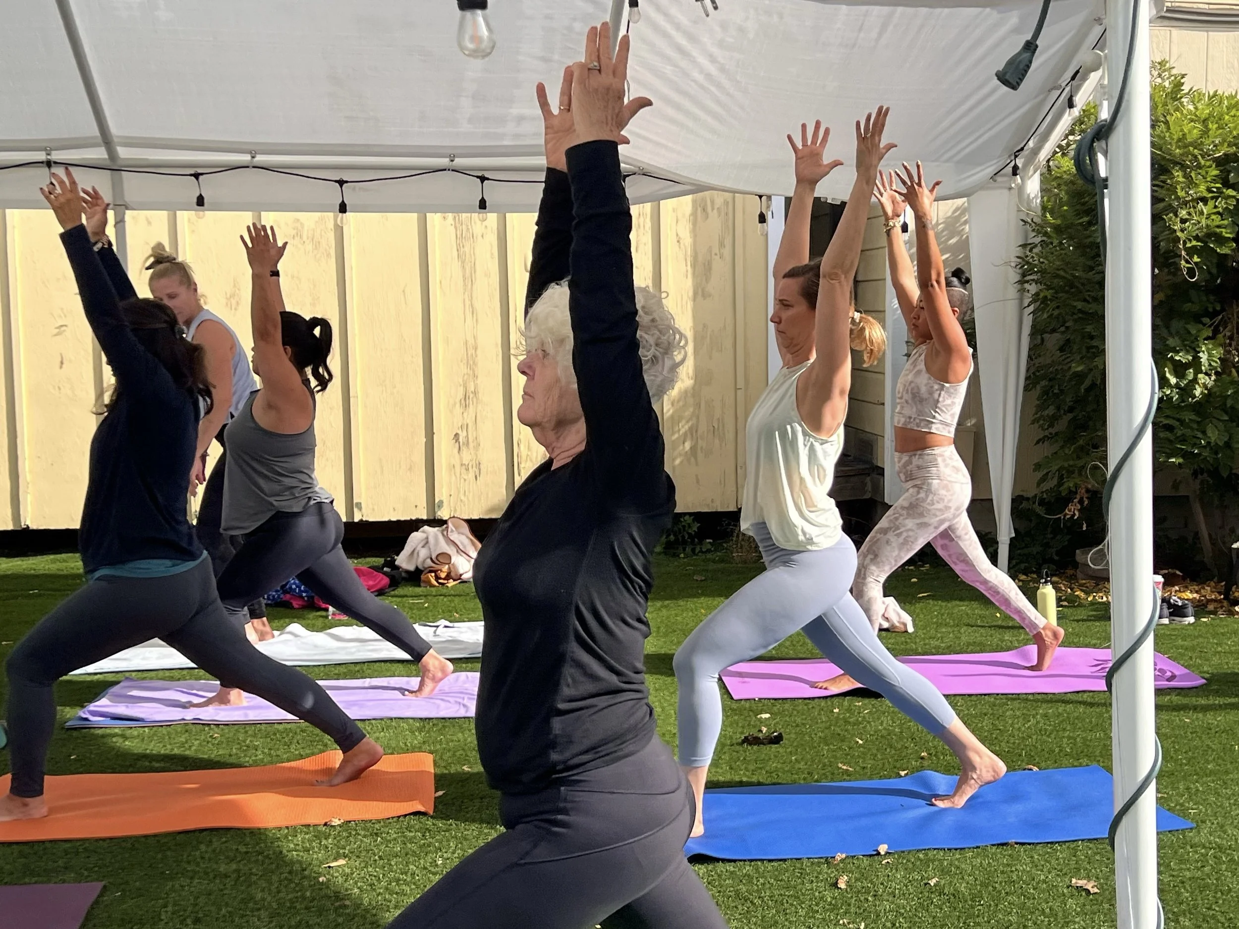 A group of women practicing yoga outdoors under a white canopy, each on a colorful yoga mat, seen in a grassy area with a wooden fence and greenery in the background.