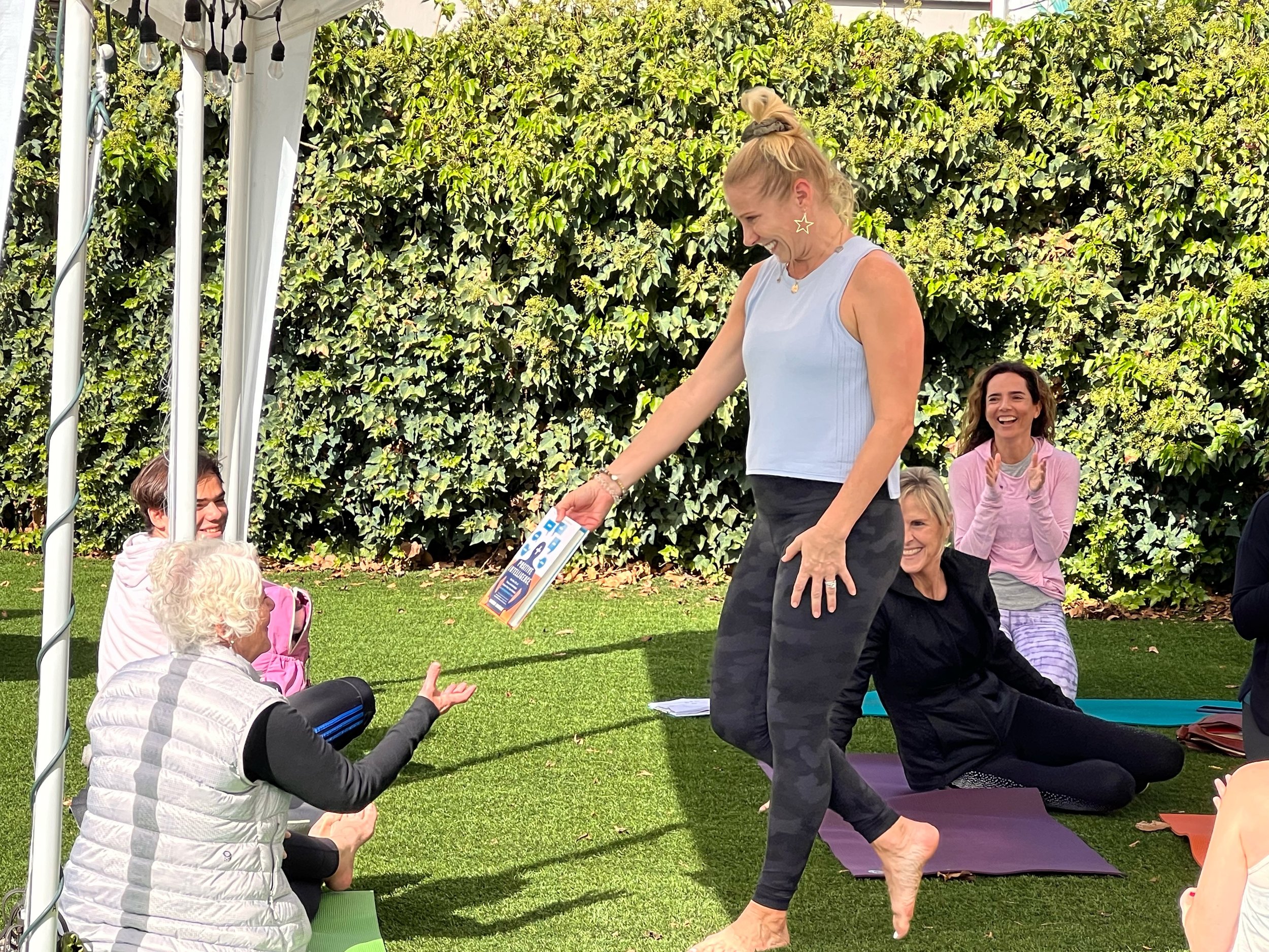 A woman wearing a light blue sleeveless top and star-shaped earrings is smiling and performing a yoga pose outdoors, surrounded by seated people clapping, with a backdrop of green foliage.