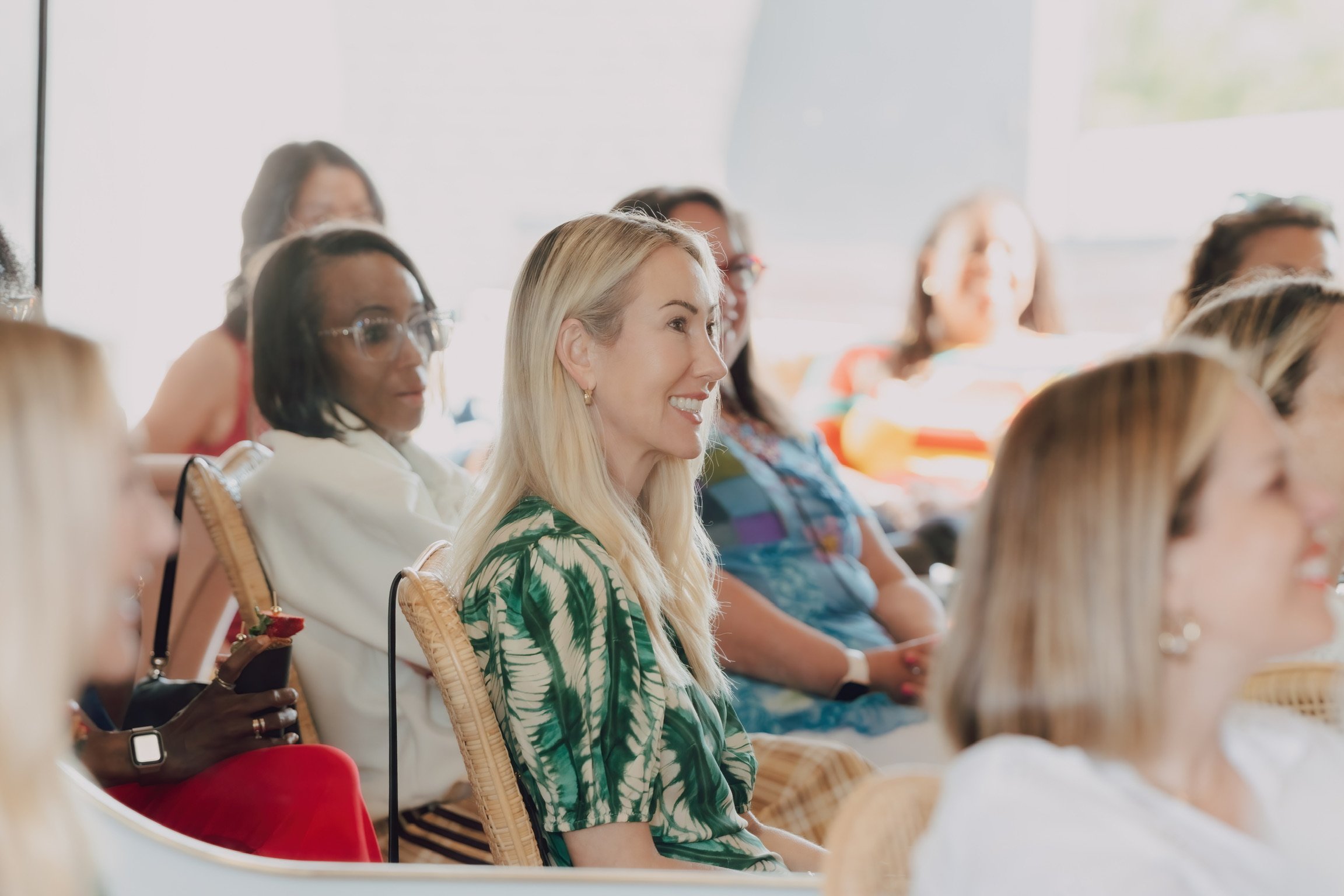A group of diverse women sitting attentively at an indoor event, with sunlight coming through large windows.