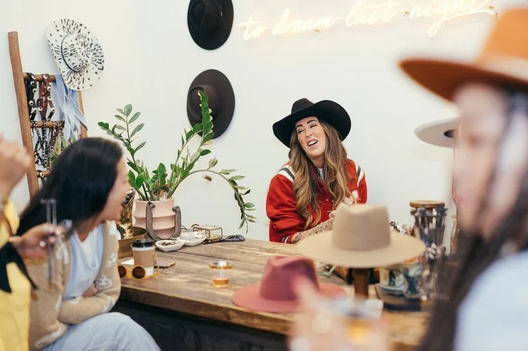 A woman with long hair wearing a black hat and red jacket is smiling and laughing at a table with friends in a cozy, stylish shop decorated with plants, hats, and wall art.