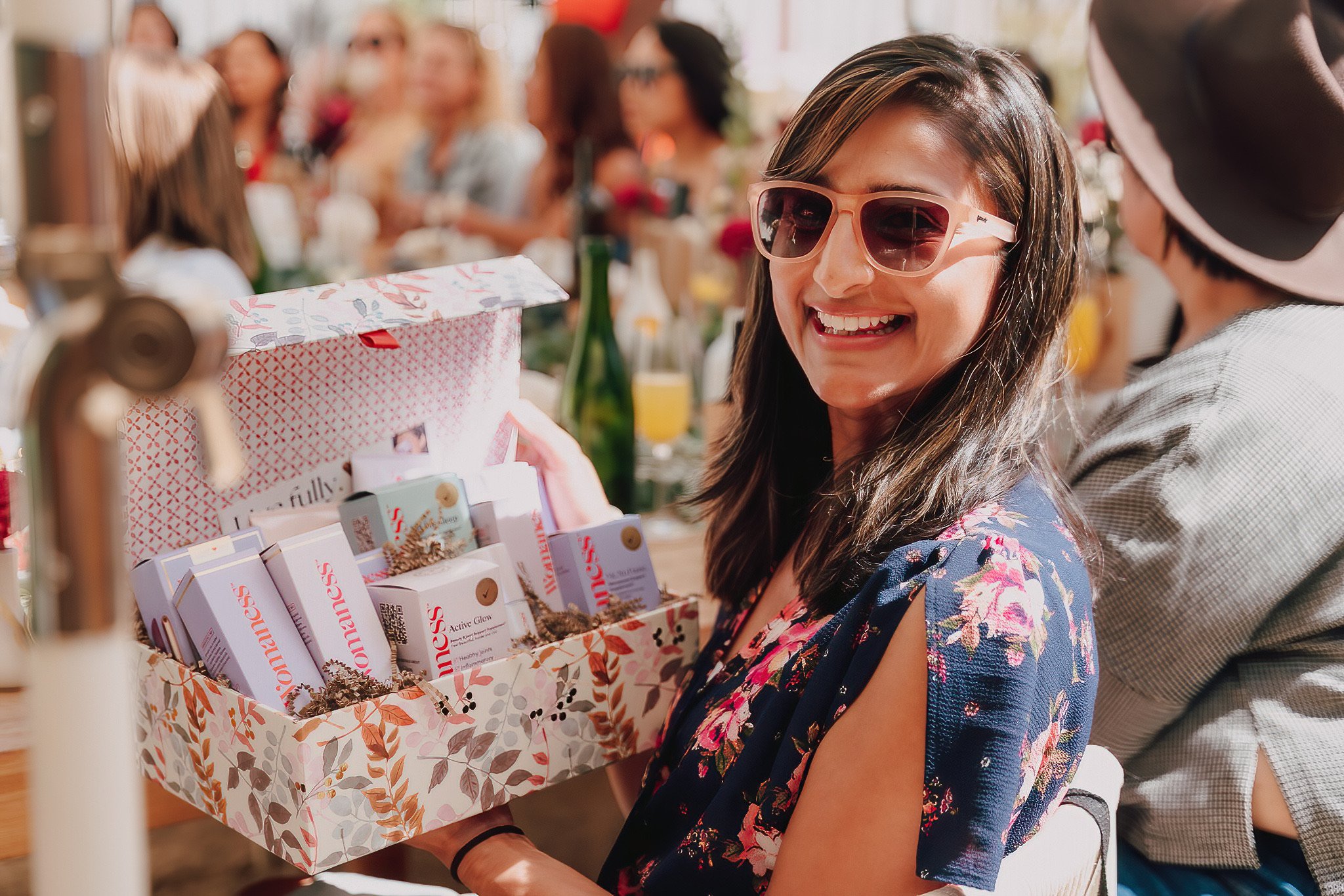 A woman with dark hair, wearing sunglasses and a floral dress, smiling as she looks at a gift box at an outdoor gathering.