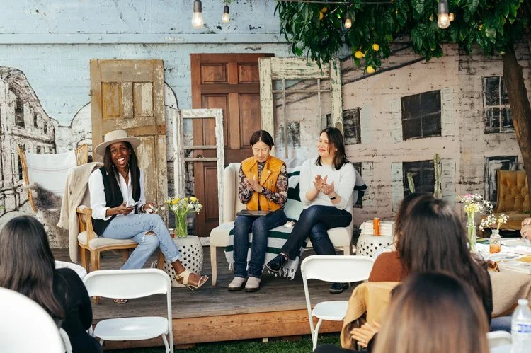 Three women sitting on a stage in front of a rustic backdrop with old doors, speaking to an audience at a casual outdoor event.