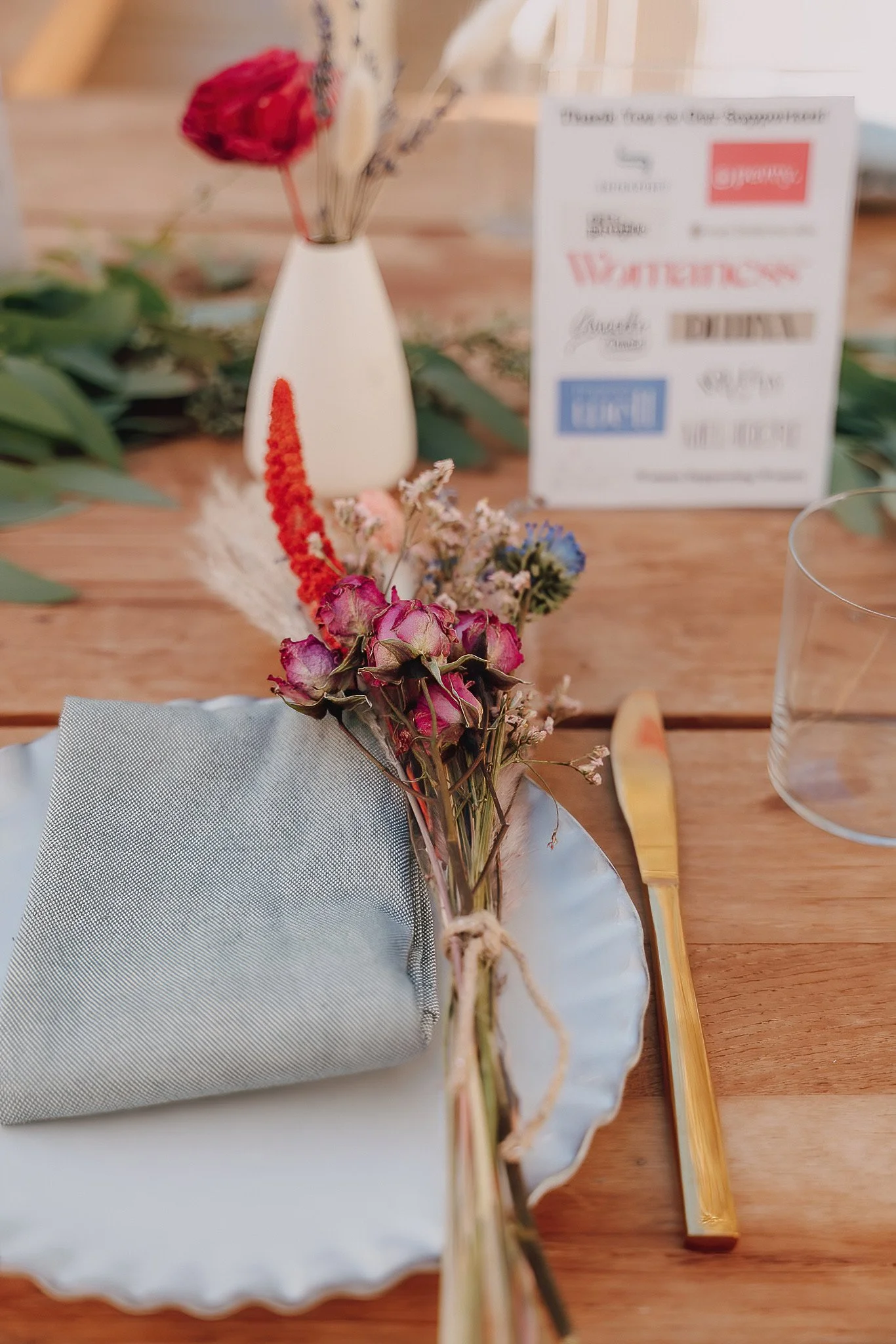Close-up of a dried floral arrangement with pink, purple, and beige flowers, a white fabric napkin on a wooden table with a clear glass and a blurred sign in the background.