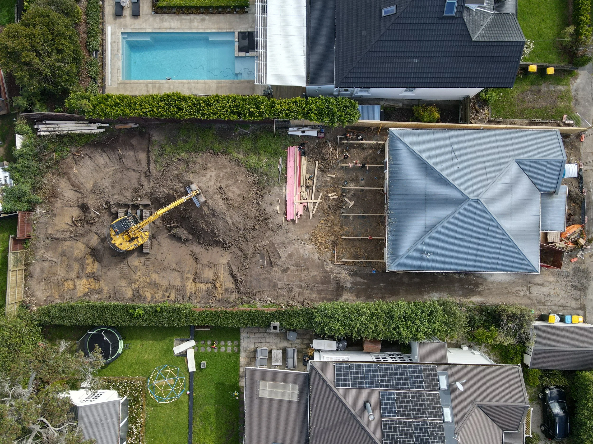 An aerial view of a backyard with a construction site, a pool, and neighboring houses. The construction area includes a small excavator and building materials.