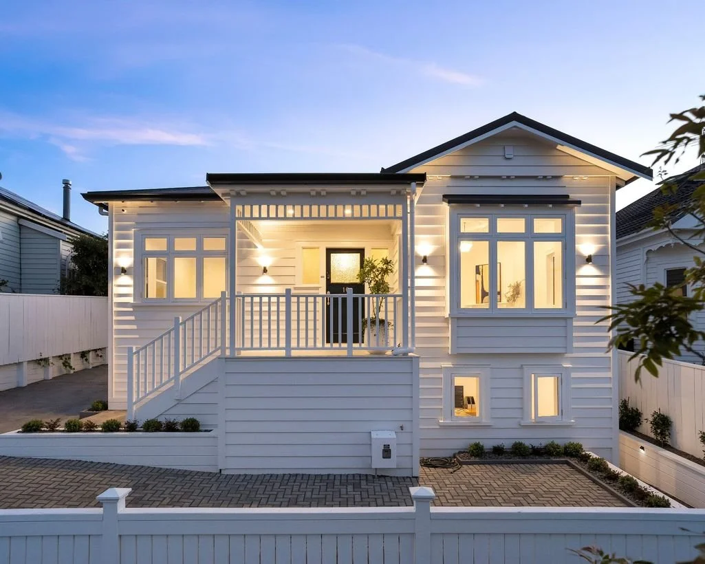 A well-lit white two-story house with a small front porch, surrounded by a white fence, during twilight with a clear sky.