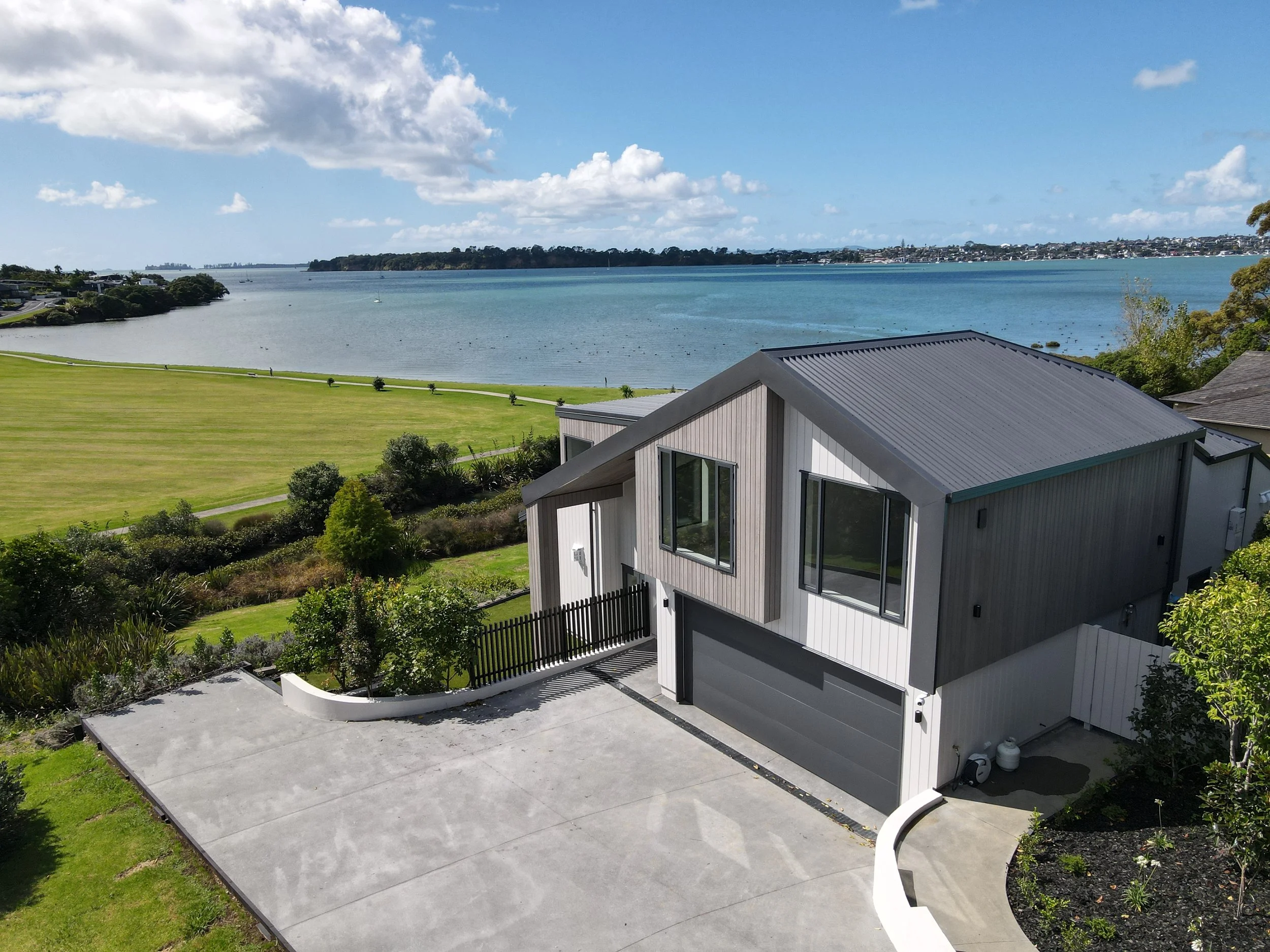A modern house with a gray metal roof and large windows, situated near a body of water with a grassy area, trees, and a distant shoreline.