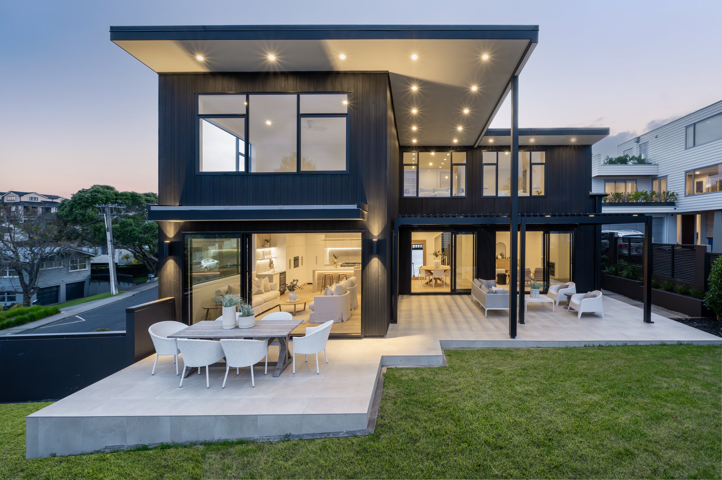 Modern two-story house with large glass windows, black exterior, and a spacious outdoor patio with white furniture and a wooden dining table, set in a landscaped yard at dusk.