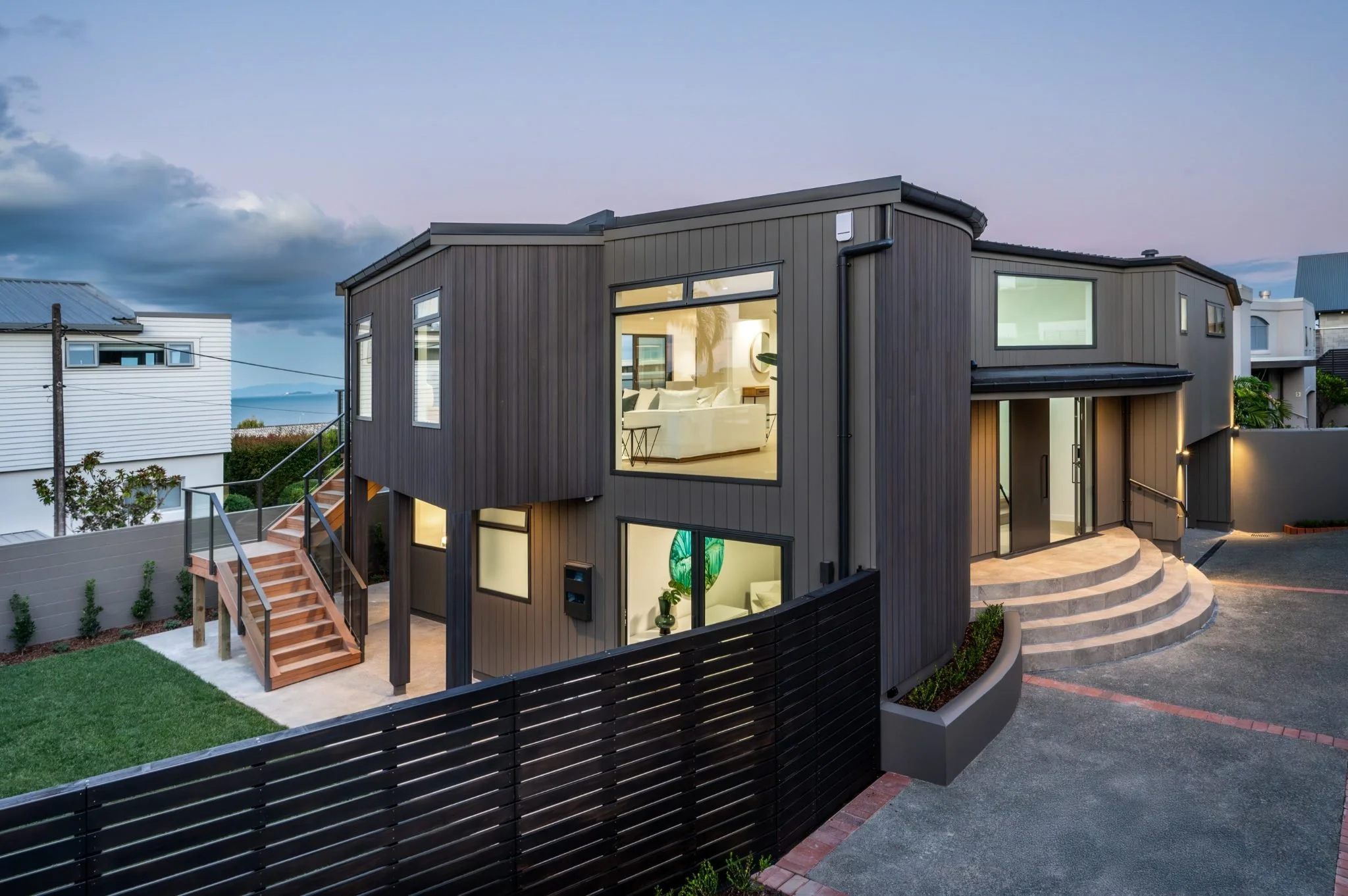 Modern two-story house with dark exterior siding, large windows, and a curved front staircase, located in a neighborhood with other houses and a view of the water in the distance.