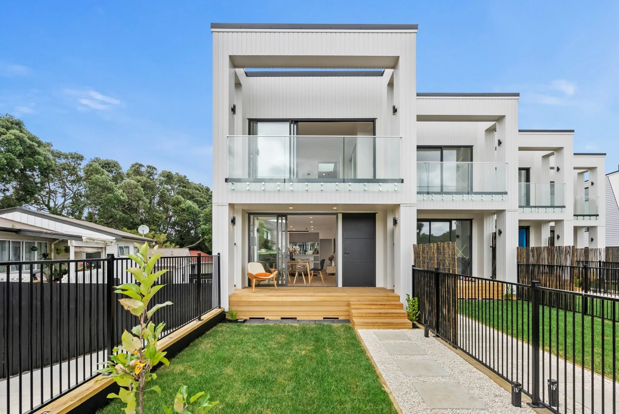 Modern multi-story white residential building with balconies and a wooden deck outdoor area, black fence, green lawn, and a clear blue sky.