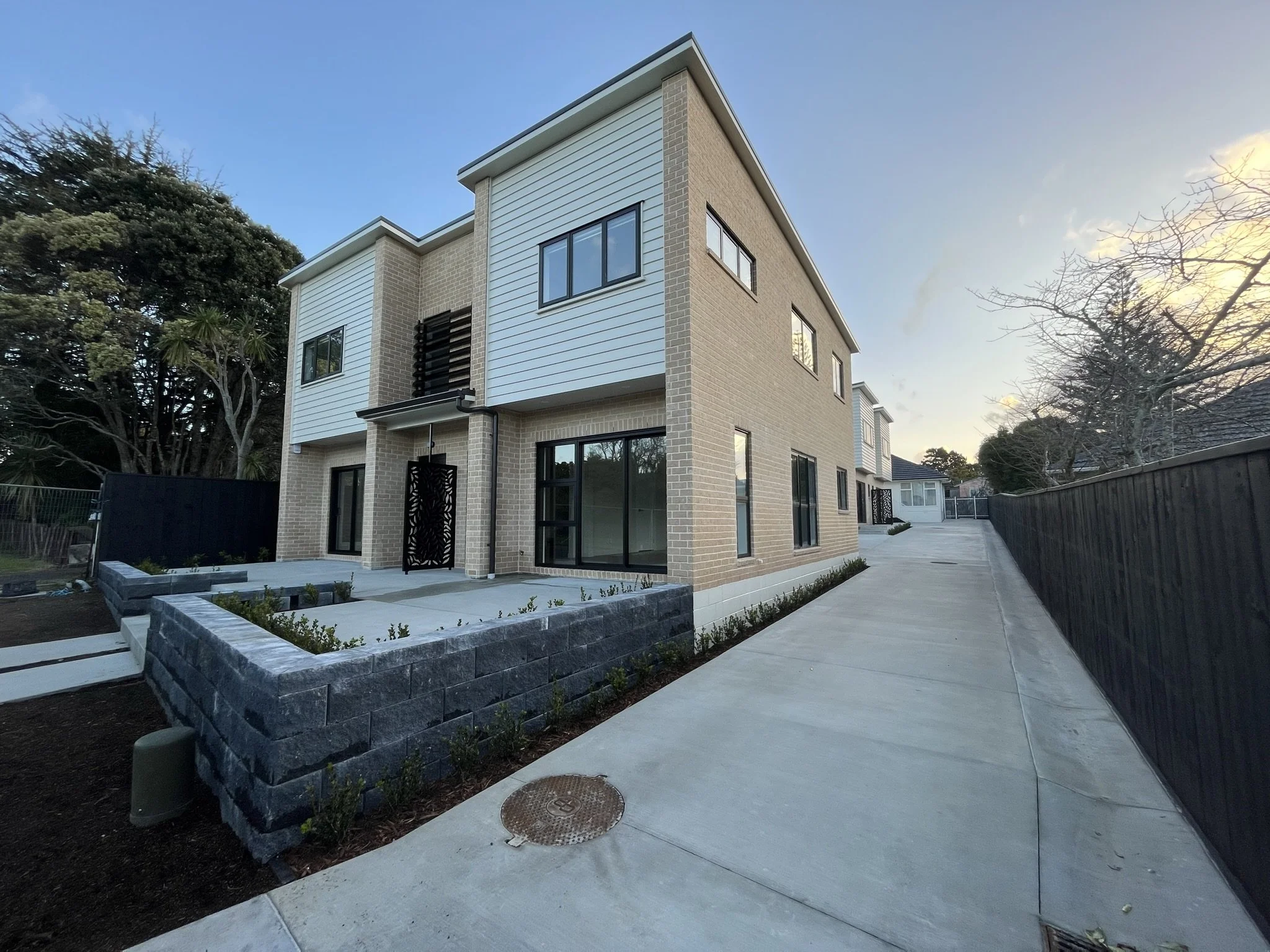 New modern multi-story residential building with brick and white siding exterior, black decorative fencing, concrete driveway, and landscaped front yard. Trees and fence in background, clear sky, and sunset lighting.