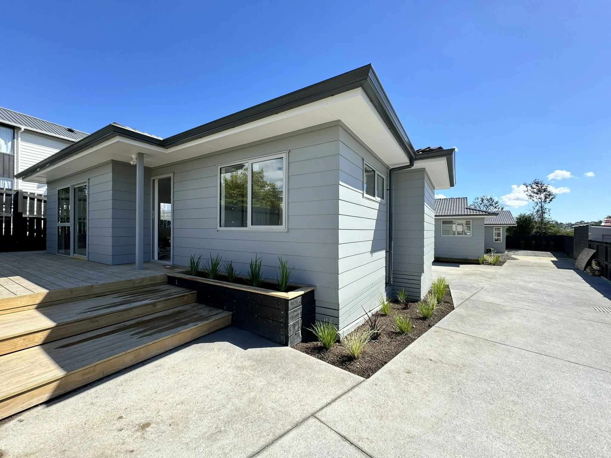 Modern house with light gray exterior, wooden stairs, and small garden with plants, under a bright blue sky.
