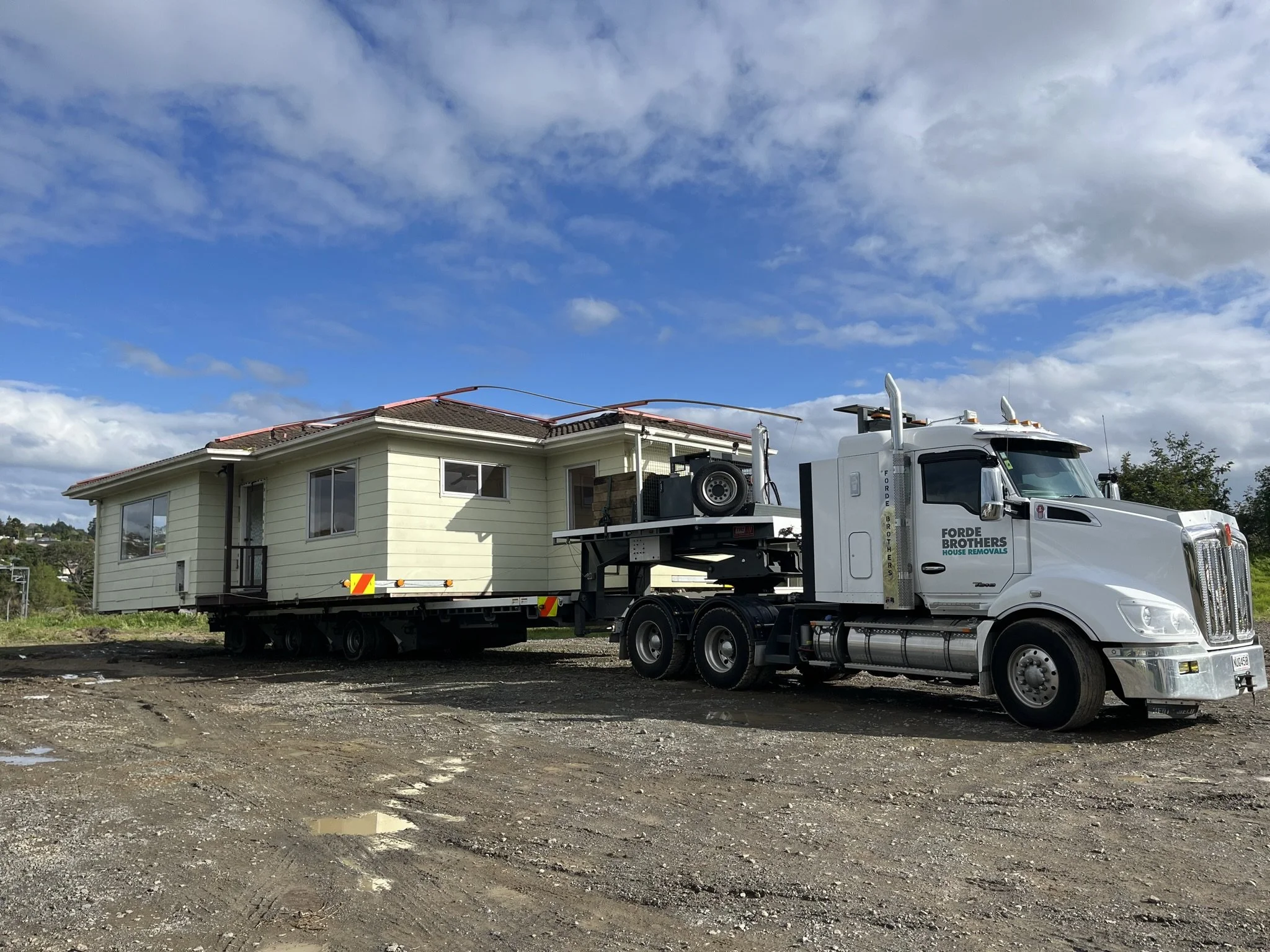 A large white moving truck labeled 'Forde Brothers House Removals' with a house on a flatbed trailer attached to the truck on a gravel lot, under a partly cloudy sky.