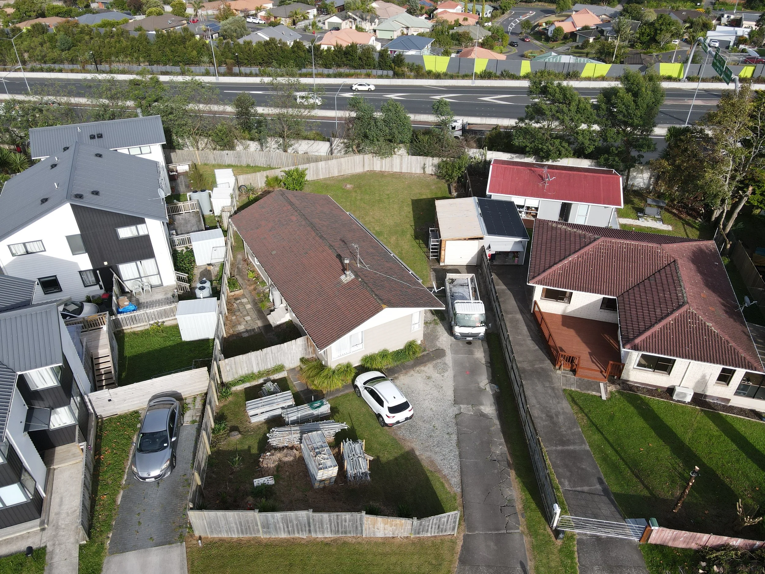 Aerial view of a neighborhood showing multiple houses with varied roof styles and colors, a driveway with parked cars, a fenced backyard with a lawn, construction materials, and a highway with a few vehicles in the background.