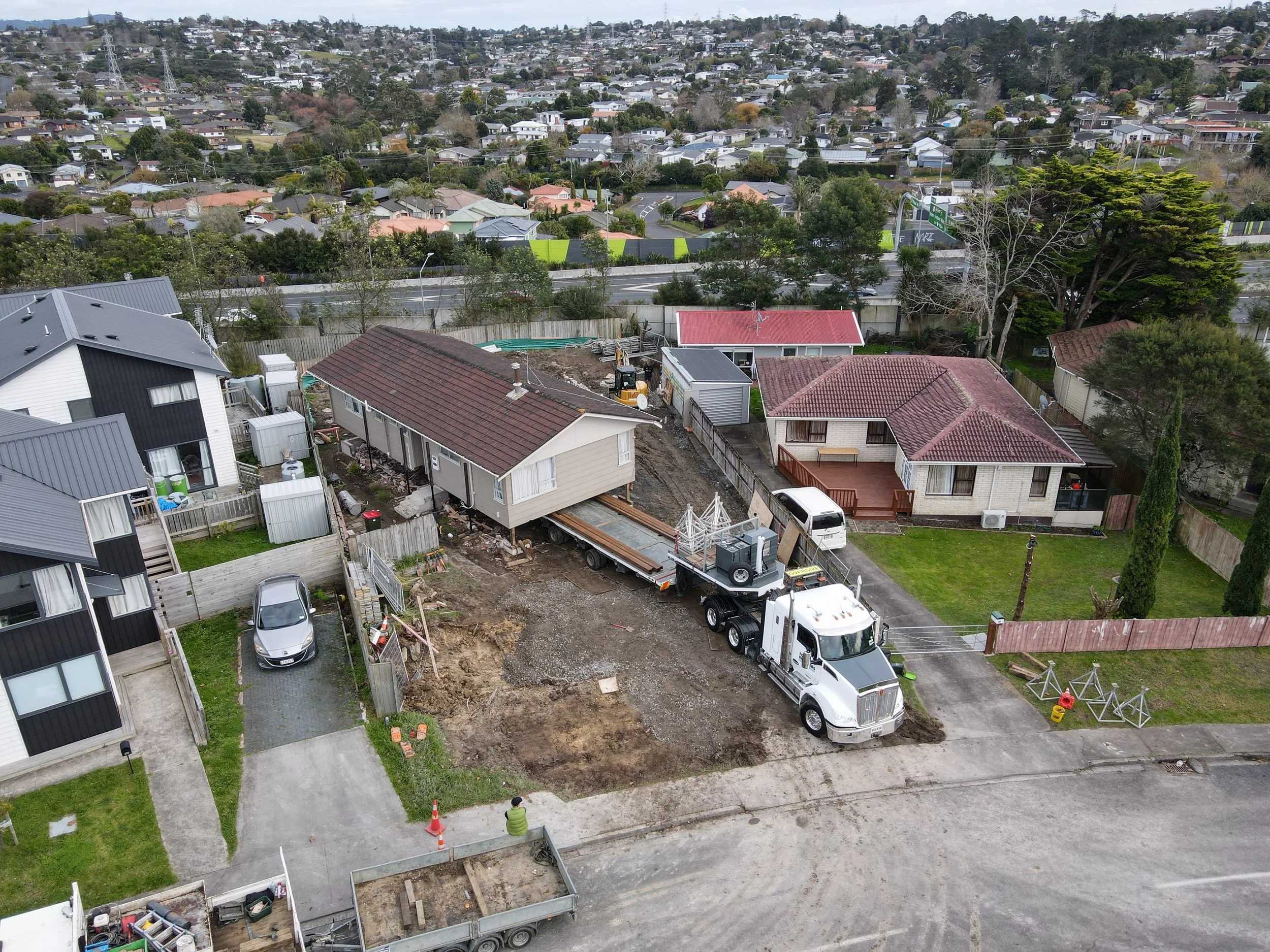Aerial view of a residential construction site with a large truck and a house on a trailer, surrounded by homes, fences, and greenery.
