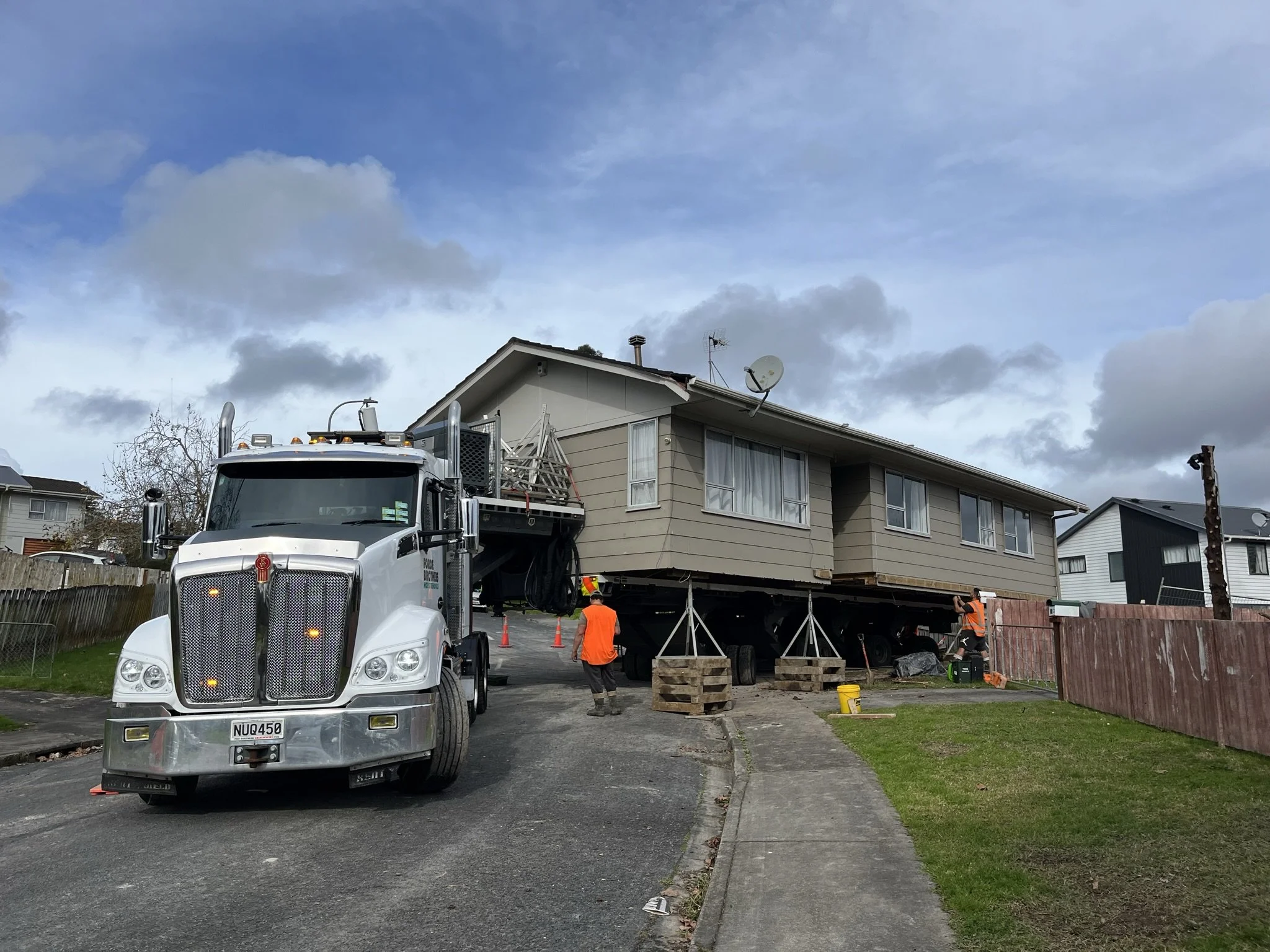 A house is being moved on a large flatbed truck, with construction workers in orange vests guiding the process on a suburban street.