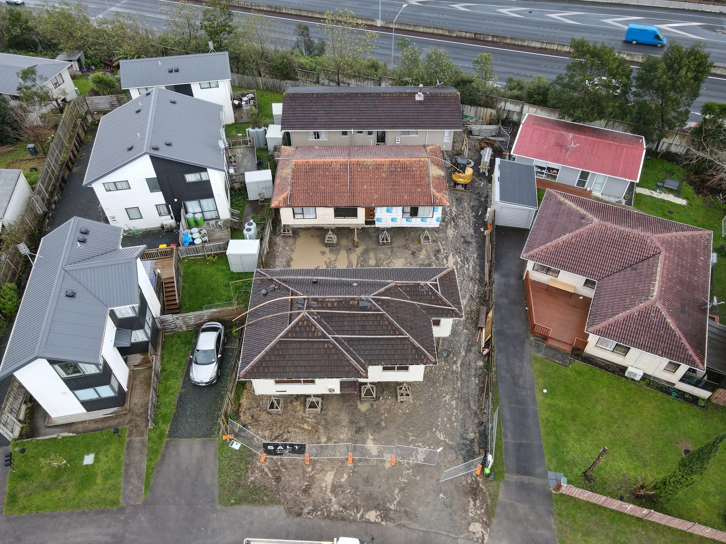 Aerial view of a residential area showing several houses, some under construction, with dirt and construction materials around. A new house is being built in the center, surrounded by other completed houses, and a highway is visible in the background.