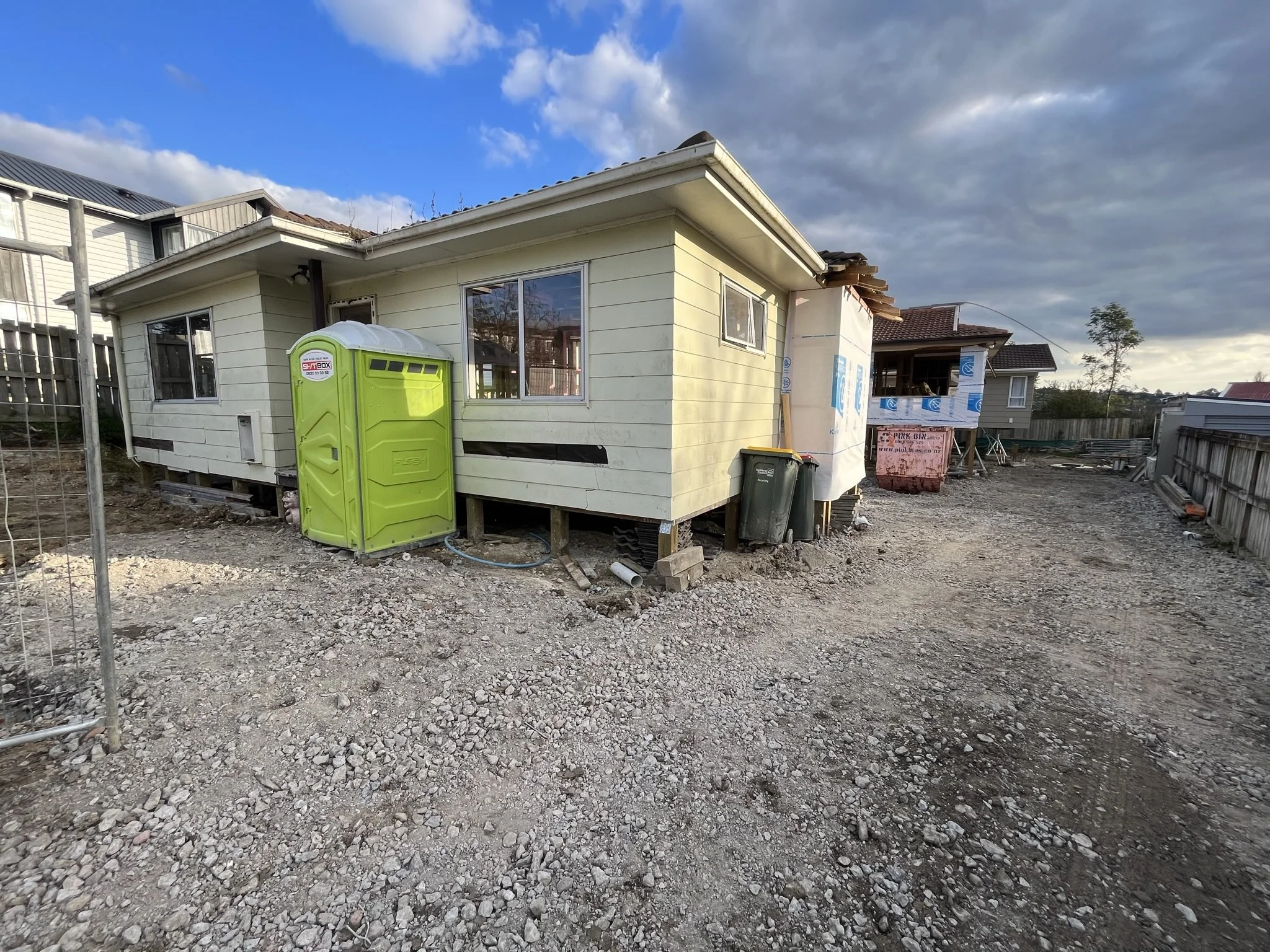View of a house under construction with siding being installed, construction materials scattered around, a bright yellow portable toilet, two trash bins, and a partly cloudy sky overhead.
