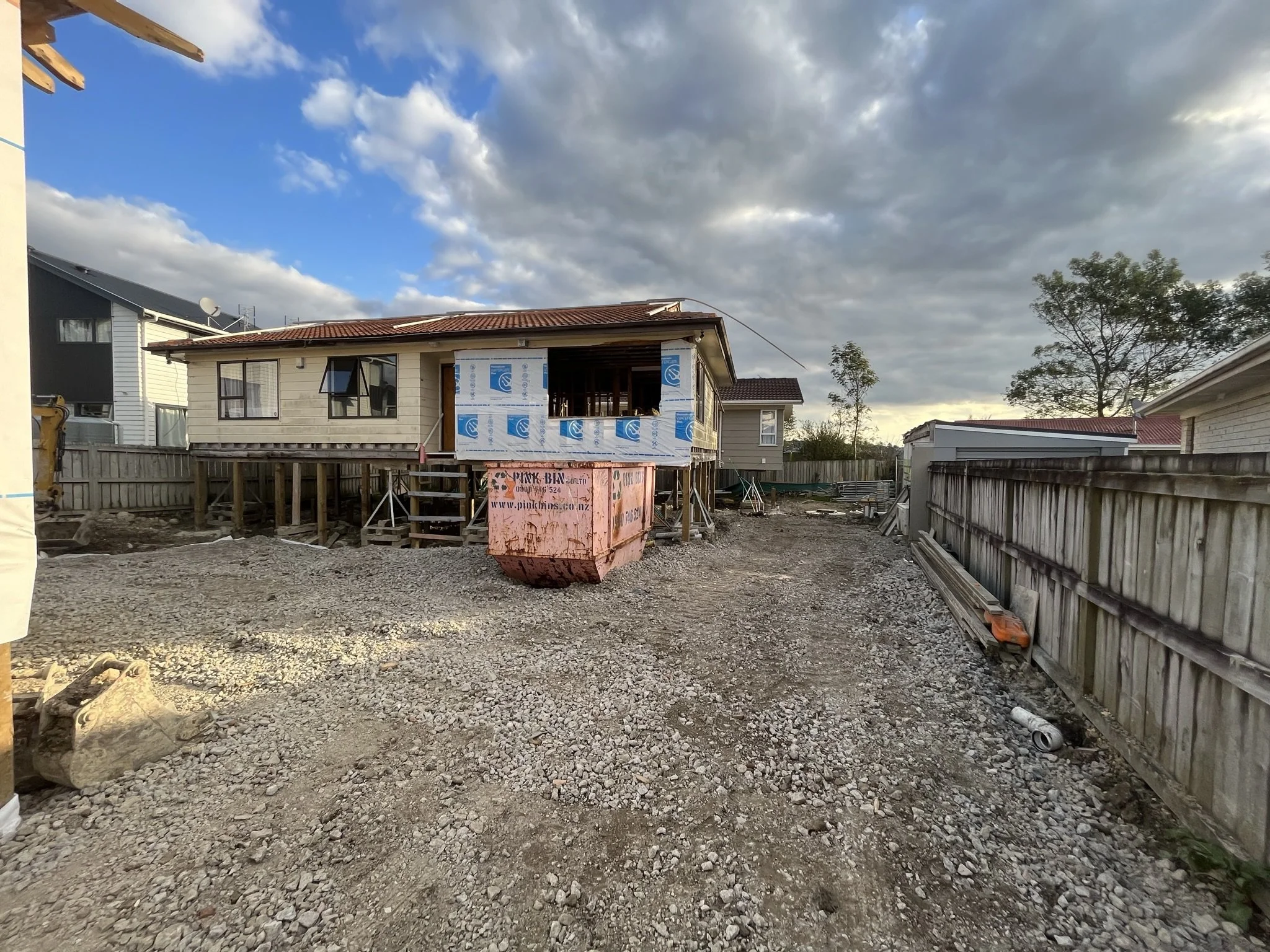 House under construction with scaffolding, wooden fence, a large pink construction dumpster, and gravel ground, against a partly cloudy sky.