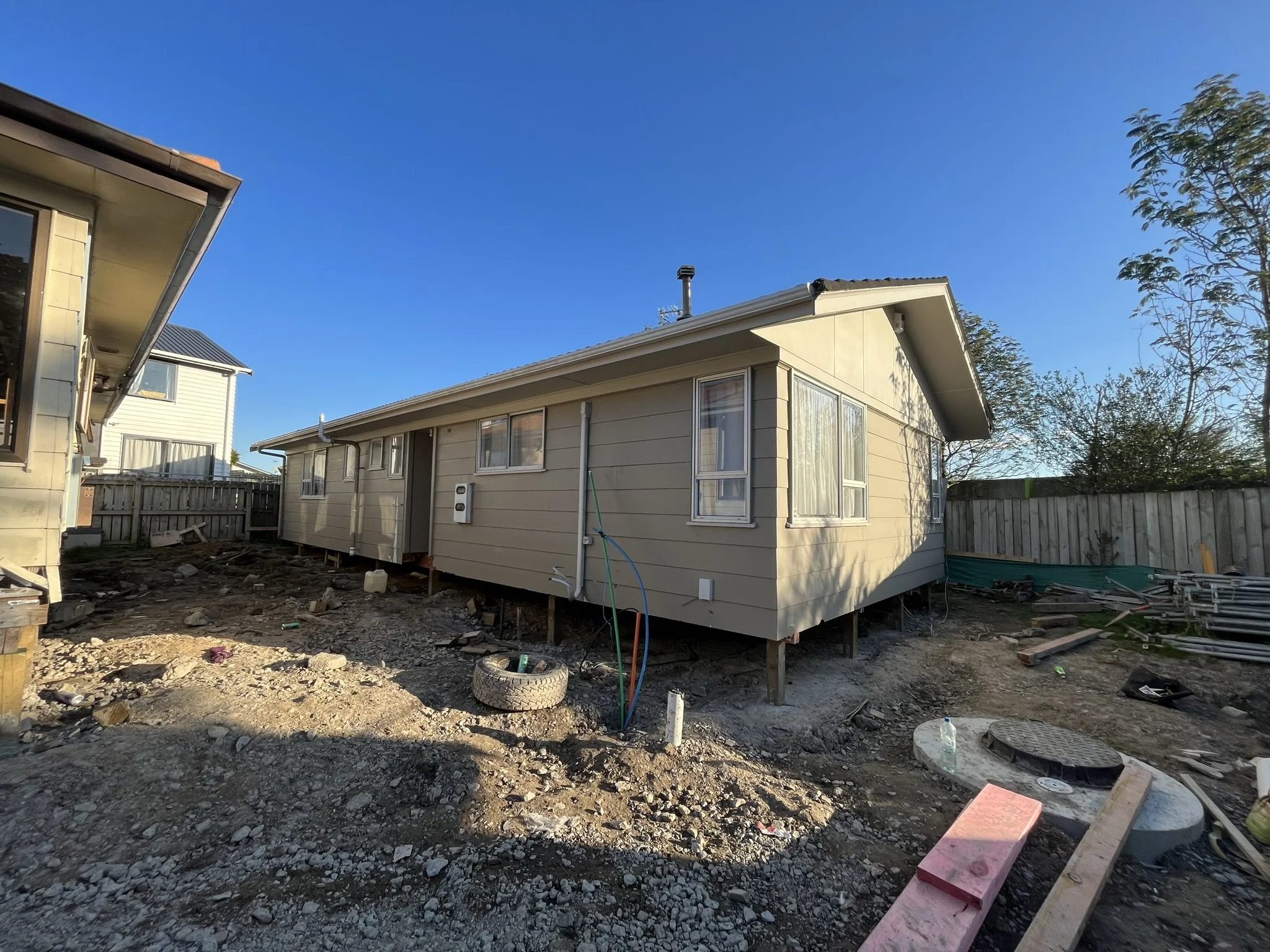A beige single-story house under construction with exposed foundation and construction debris in the yard, surrounded by wooden fences and trees, on a clear sunny day.