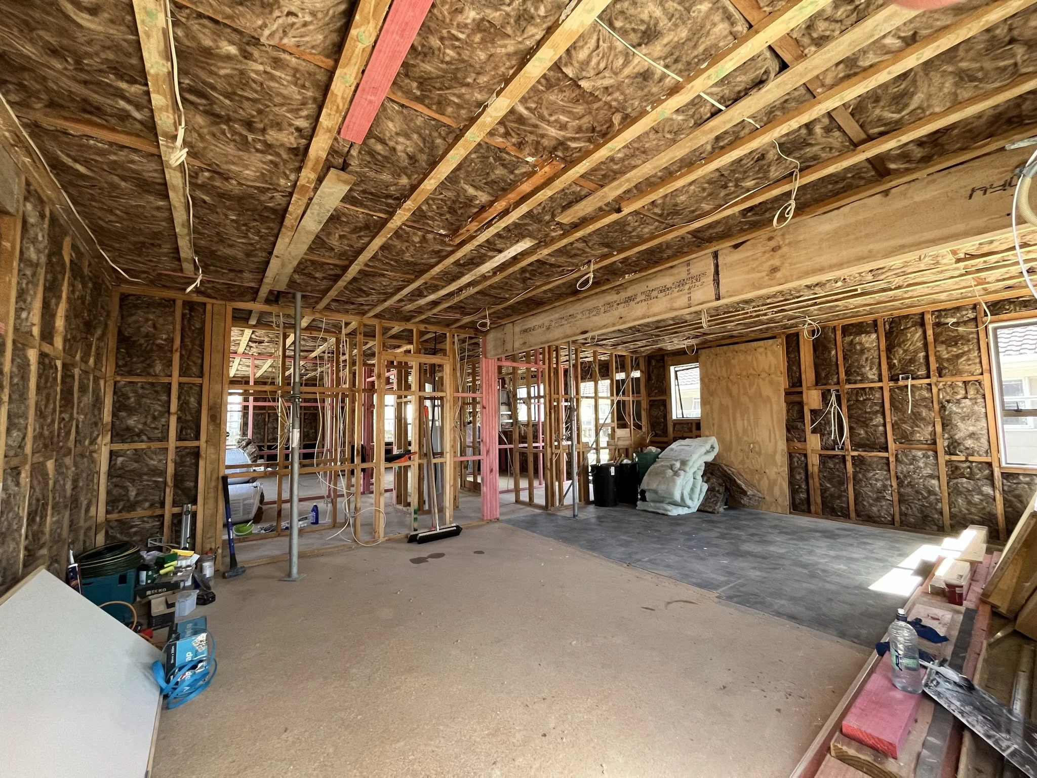 Interior of a house under construction with exposed wooden framing, insulation, and electrical wiring, and some construction tools and materials scattered around.