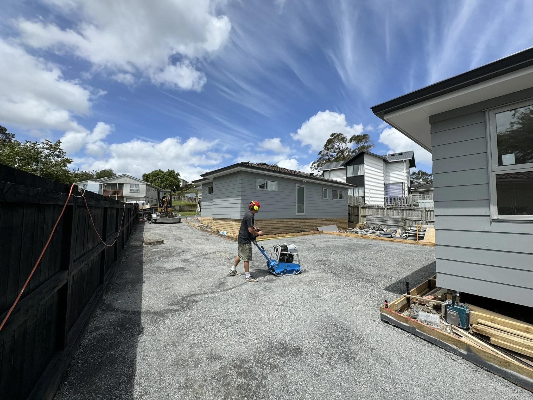 A construction worker operating a blue compactor on a gravel driveway in a residential area with houses under construction, blue sky, and scattered clouds.