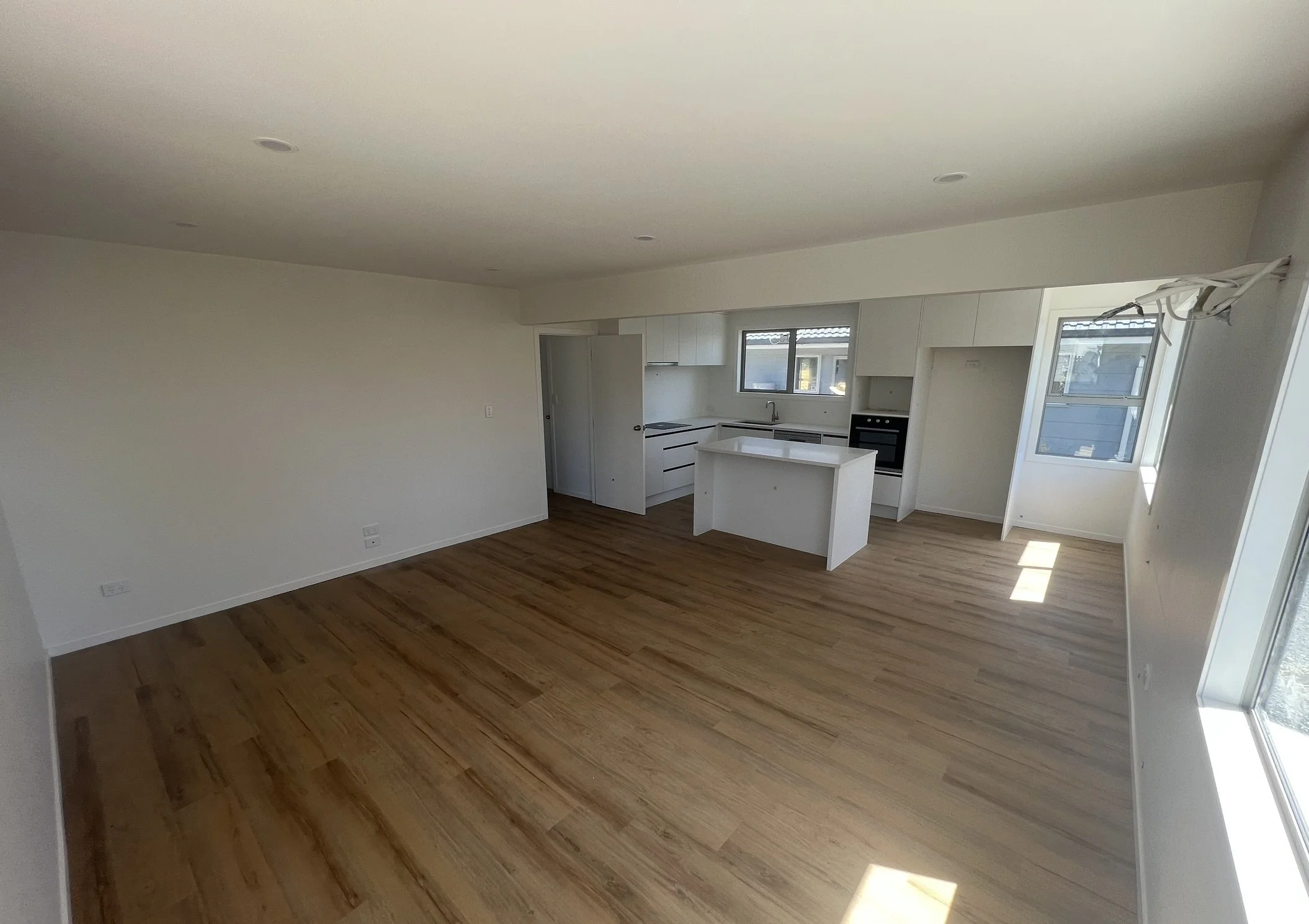 Empty living room and kitchen with white walls, hardwood floors, and large windows letting in natural light.