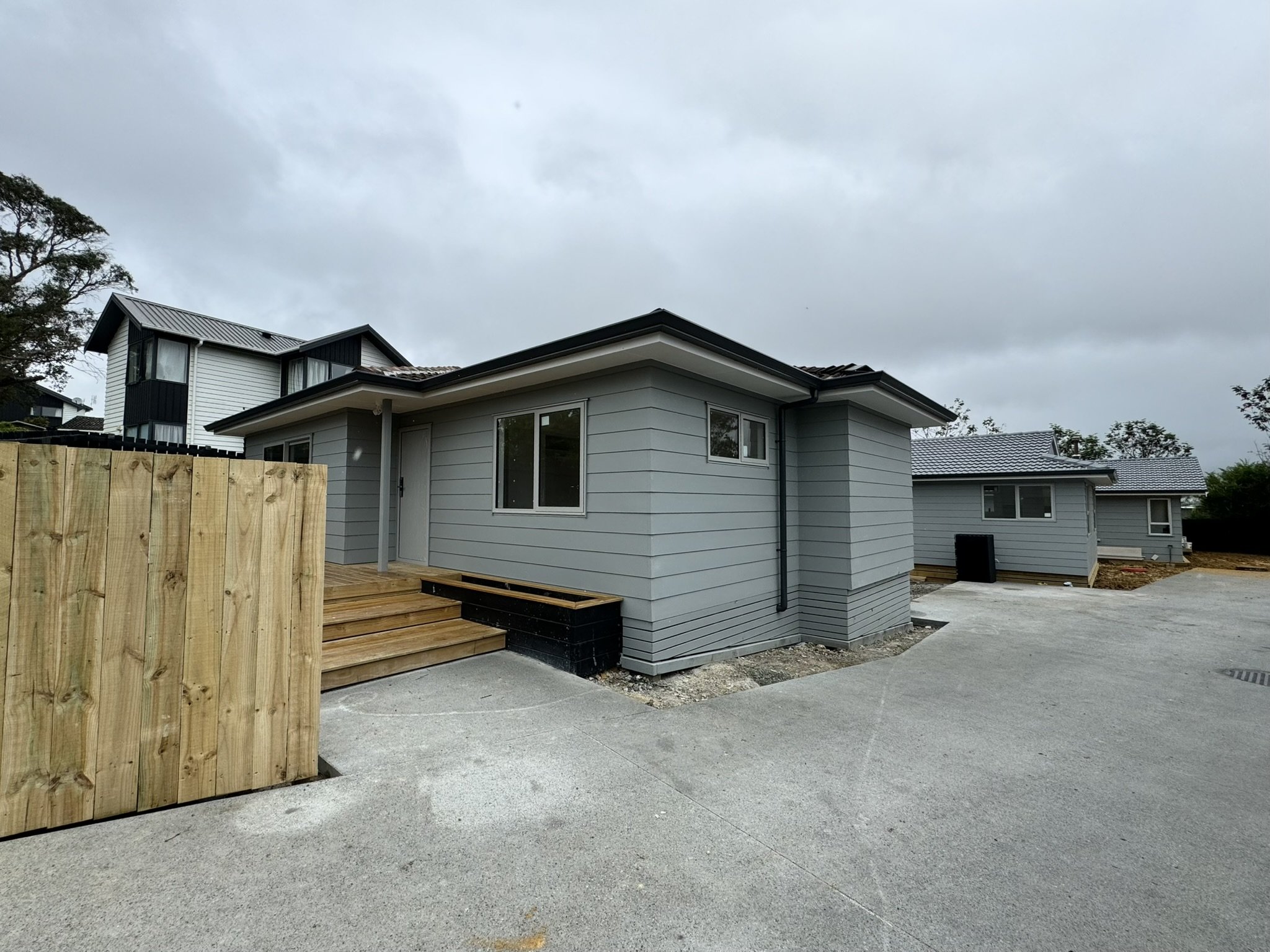 Modern house with gray siding, small front porch, and a wooden staircase, surrounded by concrete driveway and construction area under a cloudy sky.