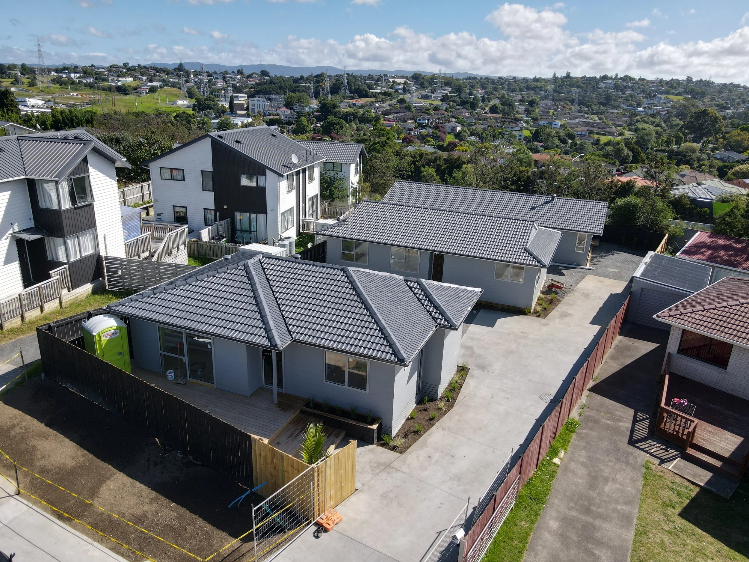 Aerial view of a new residential neighborhood with several modern houses featuring gray tiled roofs, fenced yards, some with small decks, surrounded by greenery and distant hills under a partly cloudy sky.