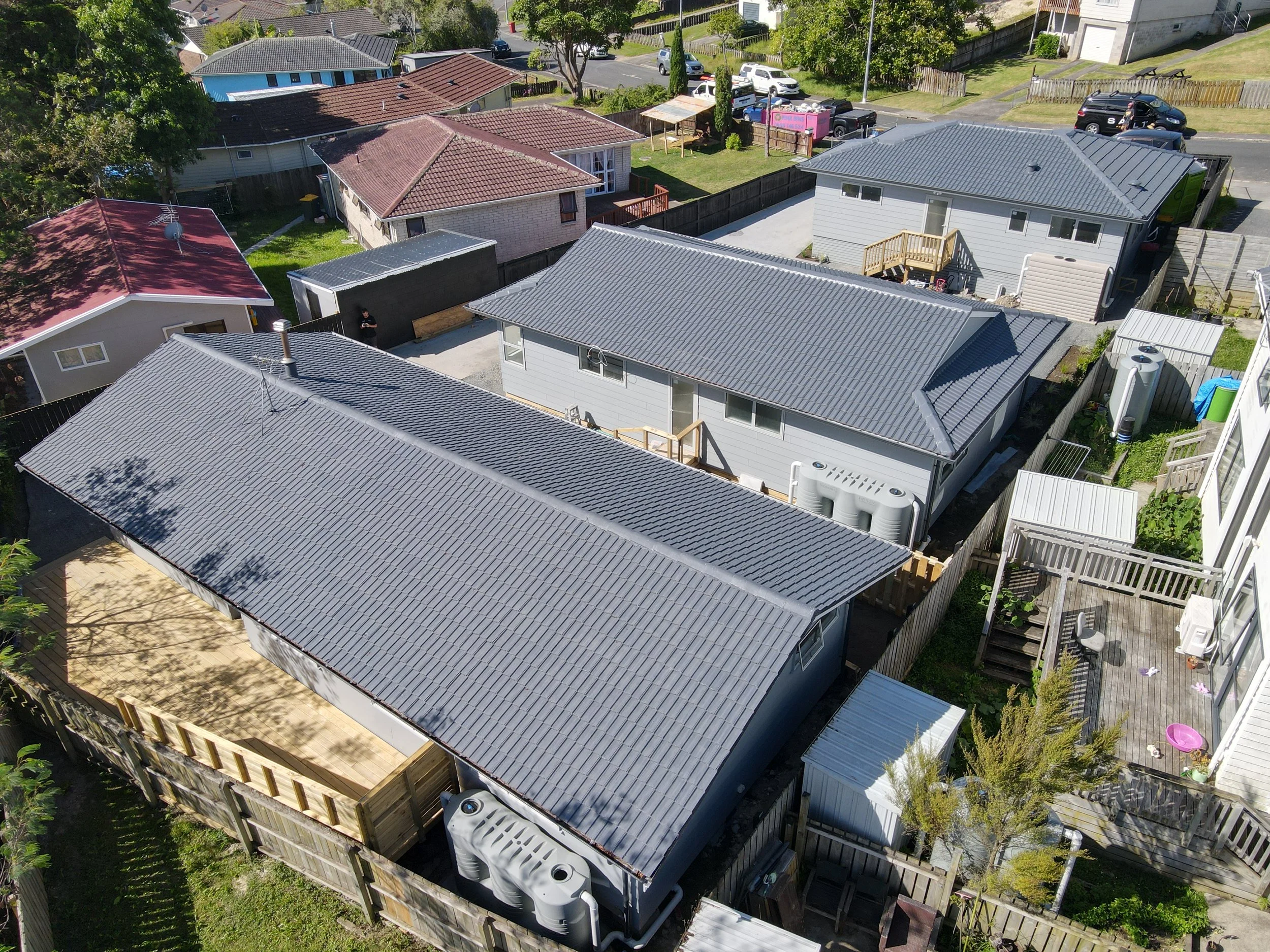 Aerial view of residential neighborhood with multiple houses and backyards, featuring gray metal roofs, fenced yards, trees, and some parked cars on the street.