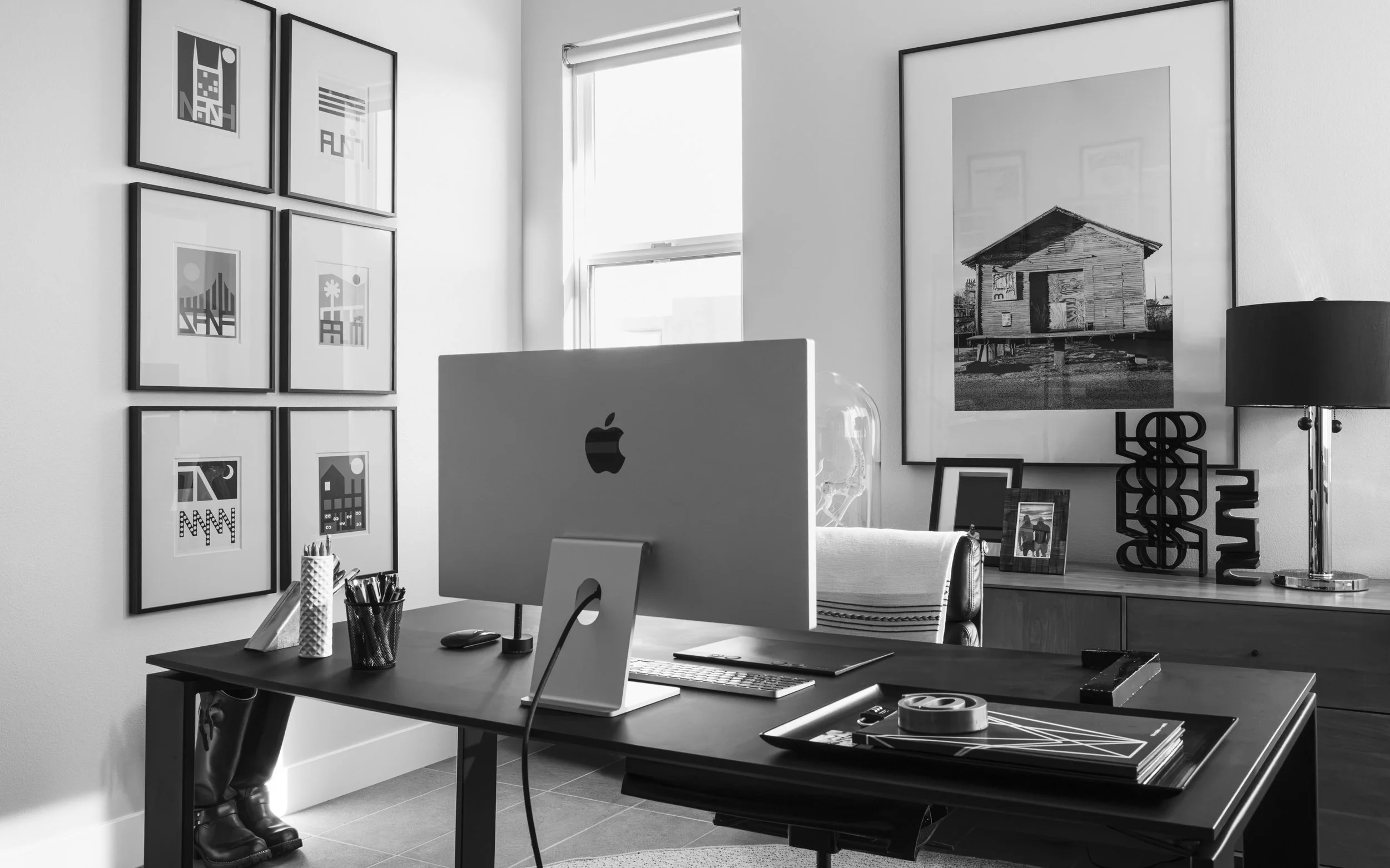 A modern home office featuring a desk with an iMac computer, various office supplies, and a large framed black-and-white photograph of a house on the wall. The room is well-lit by natural light coming through a window with blinds. On the left wall, there are nine framed abstract art pieces arranged in a grid. There is a table with decorative items, including a lamp and framed photos, in the background.