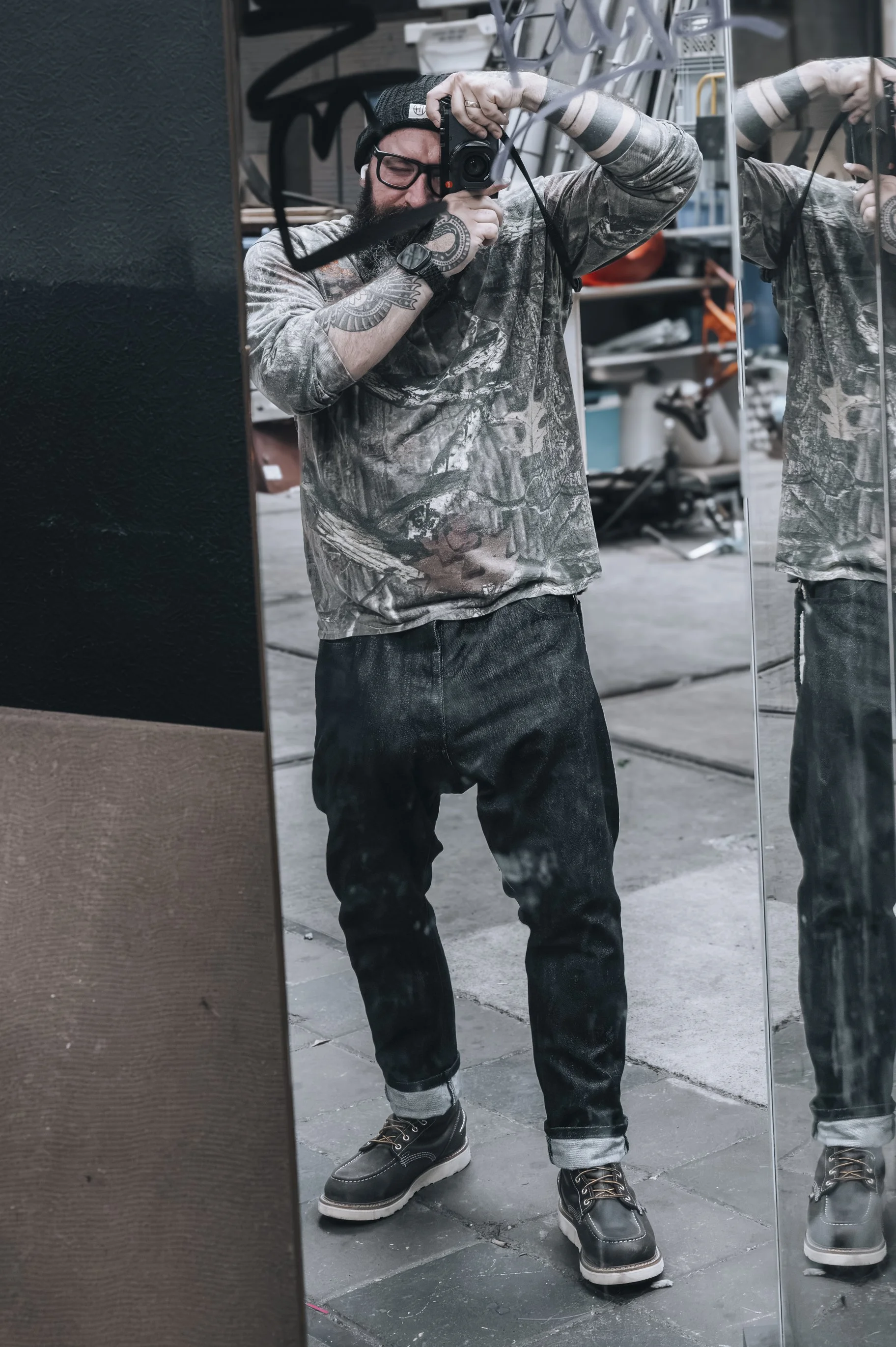 Sean Lopano man taking a selfie in front of a mirror in a workshop or garage, wearing a camouflage sweatshirt, black pants, and boots, with tattoos on his arm, a beanie, and glasses. Taken at the NDSM complex in Amsterdam.