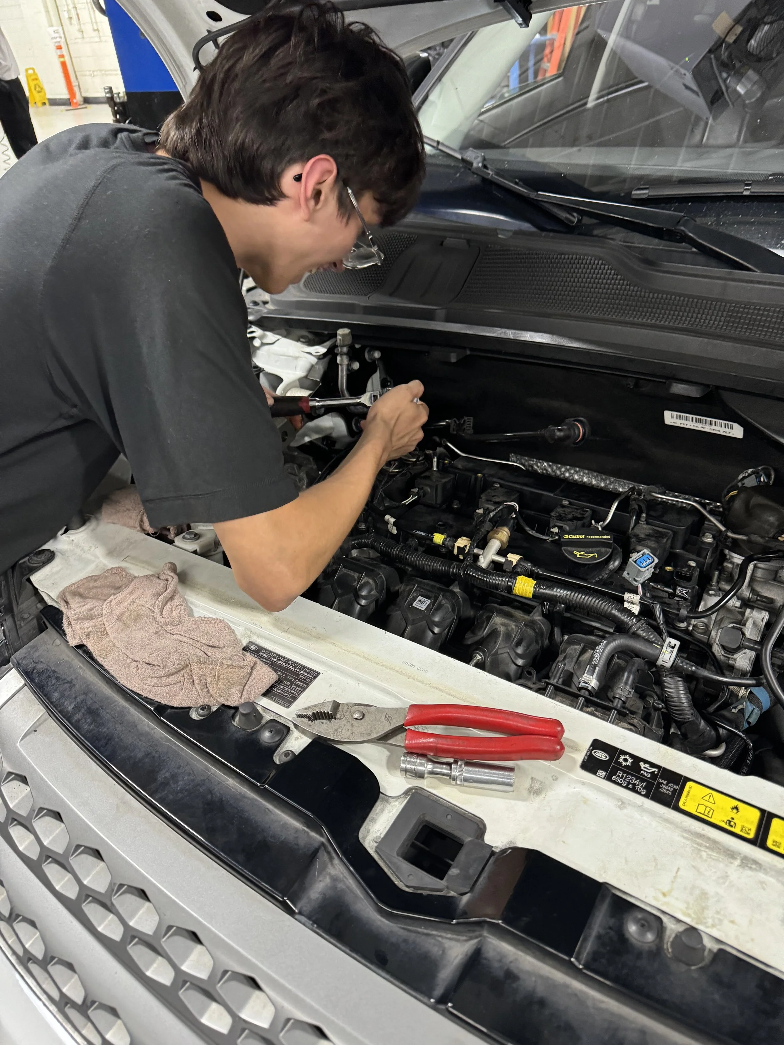 A person working on a car engine in an auto repair shop. The person is using tools near the engine with the car's hood open. A cloth and pliers are visible on the front of the car.