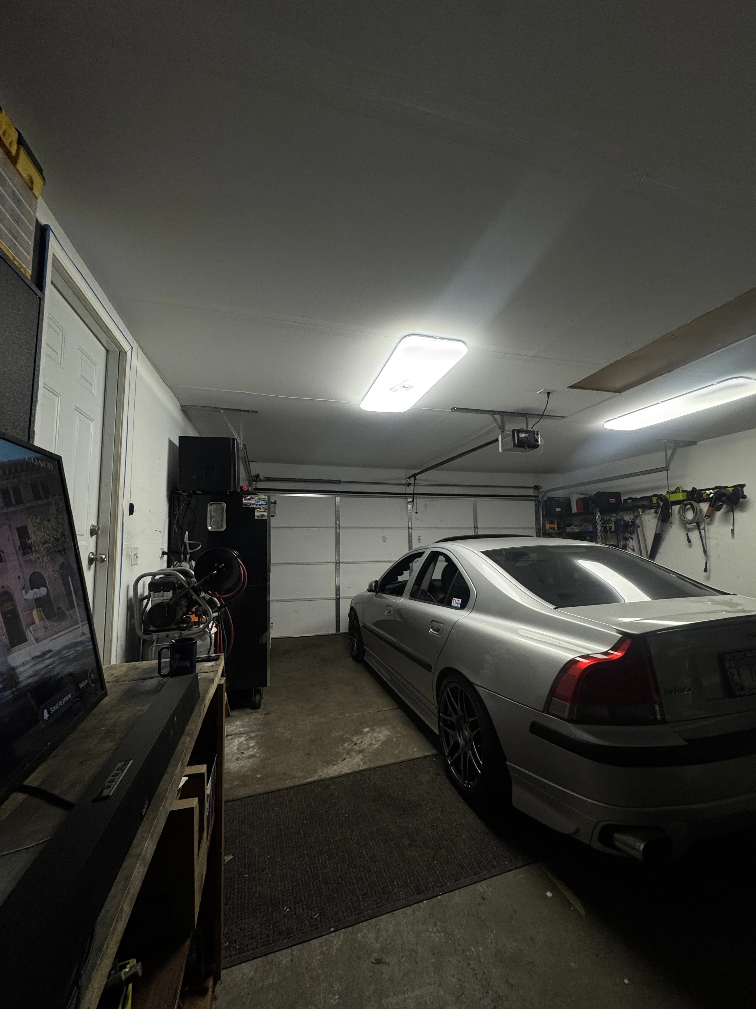 A silver sports car parked inside a garage with storage shelves and tools on the side wall. The garage has a white ceiling with fluorescent lighting and a concrete floor.