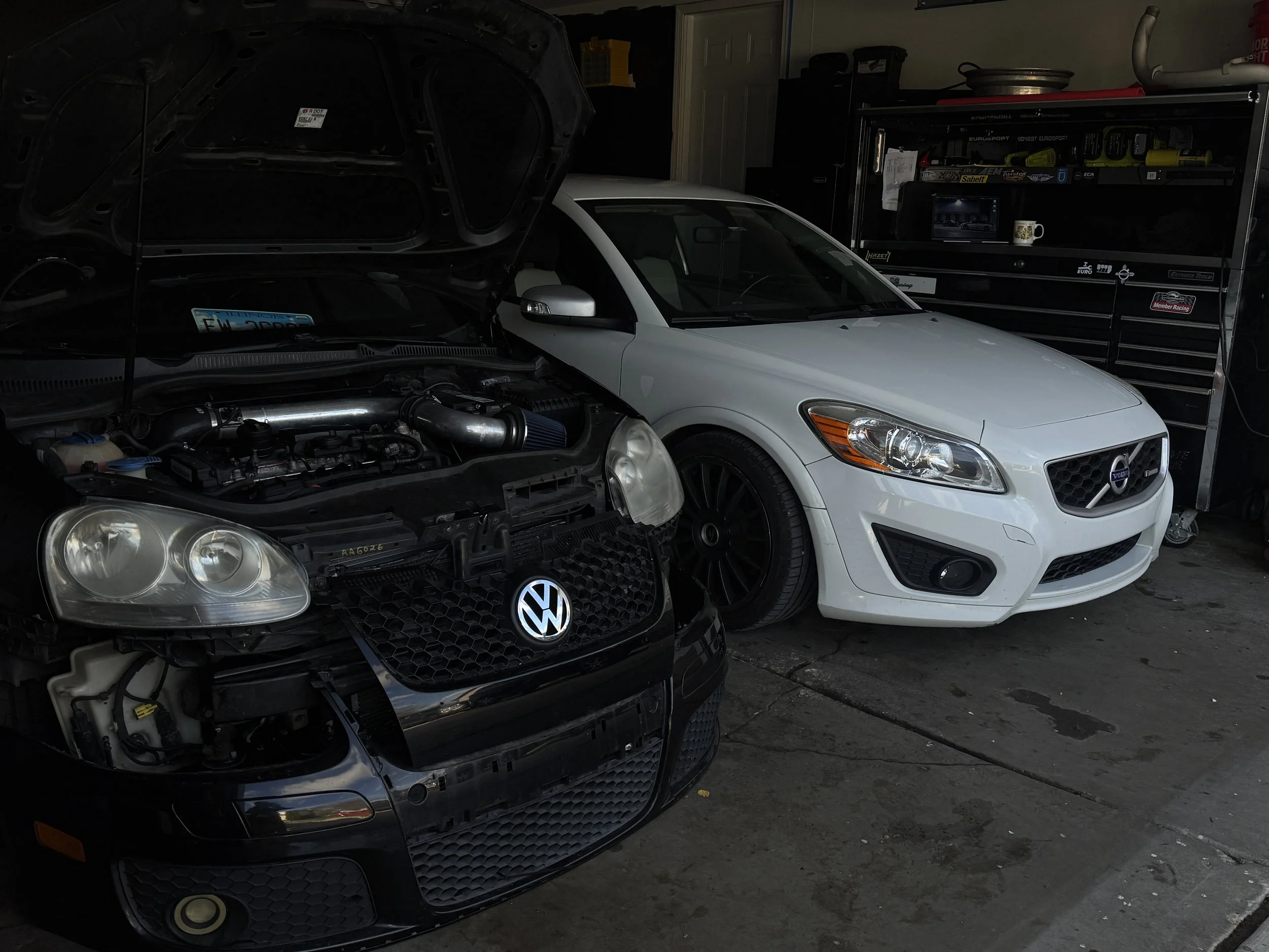 A partially disassembled black Volkswagen car with its hood open, showing the engine bay, parked next to a white car in a garage. A toolbox with various tools is visible in the background.