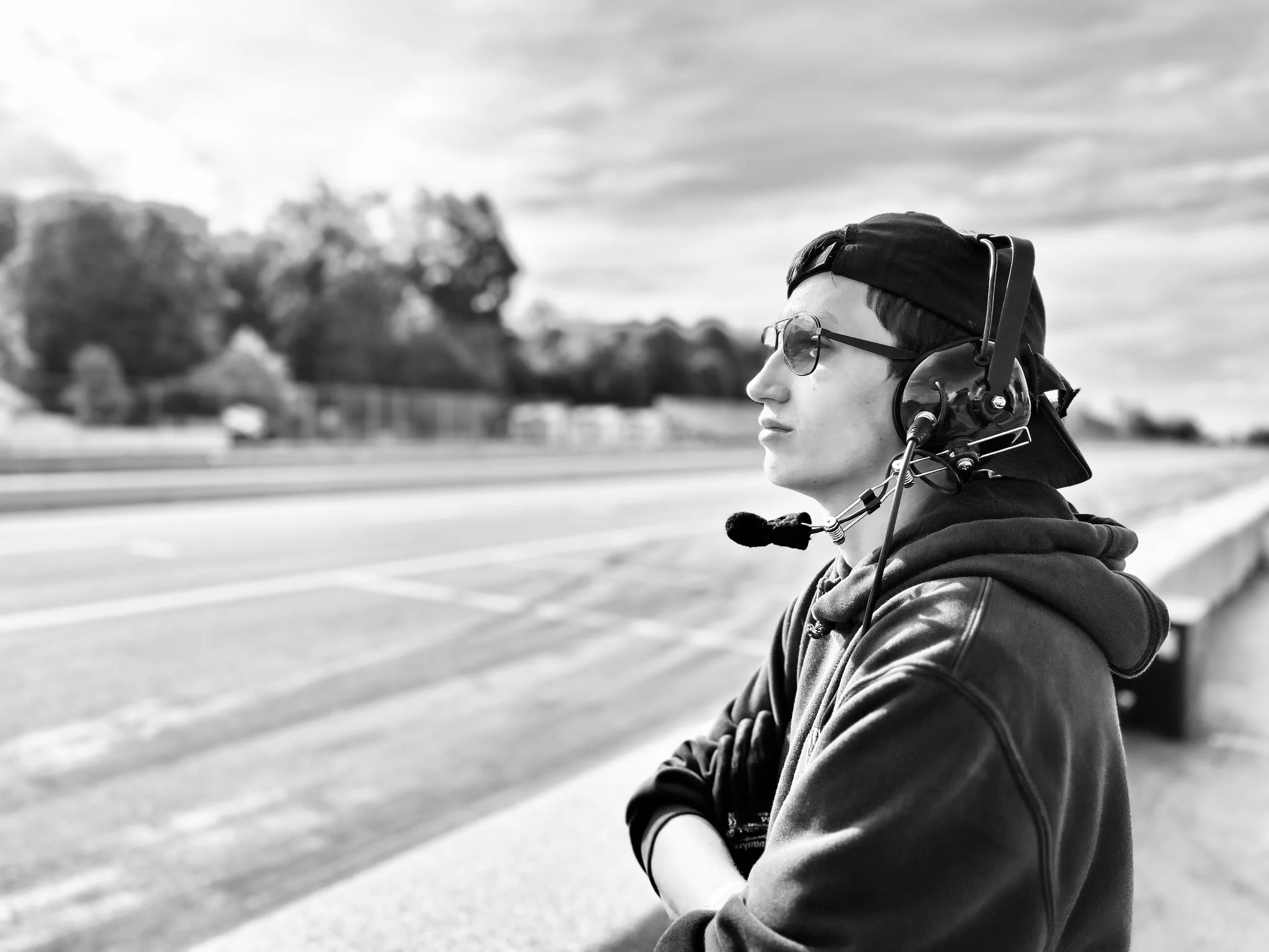 A young person wearing glasses, a baseball cap backwards, and a headset with a microphone, sitting outdoors and looking to the right.