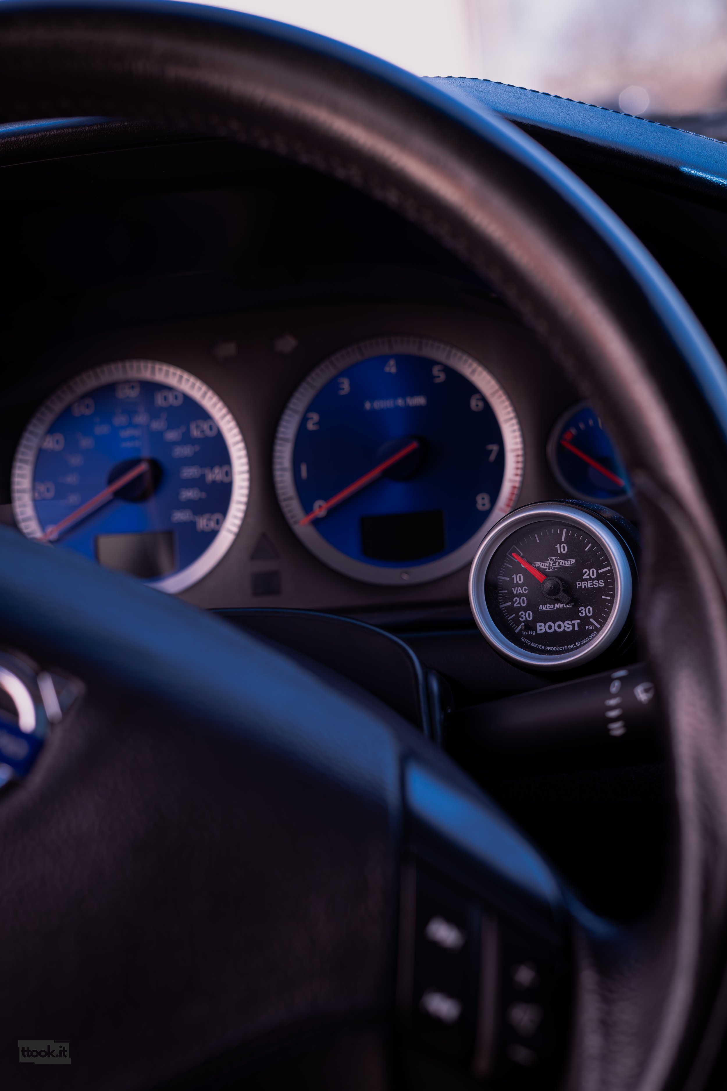 Close-up of a car dashboard showing speedometer, tachometer, and an aftermarket boost gauge, with part of the steering wheel in view.