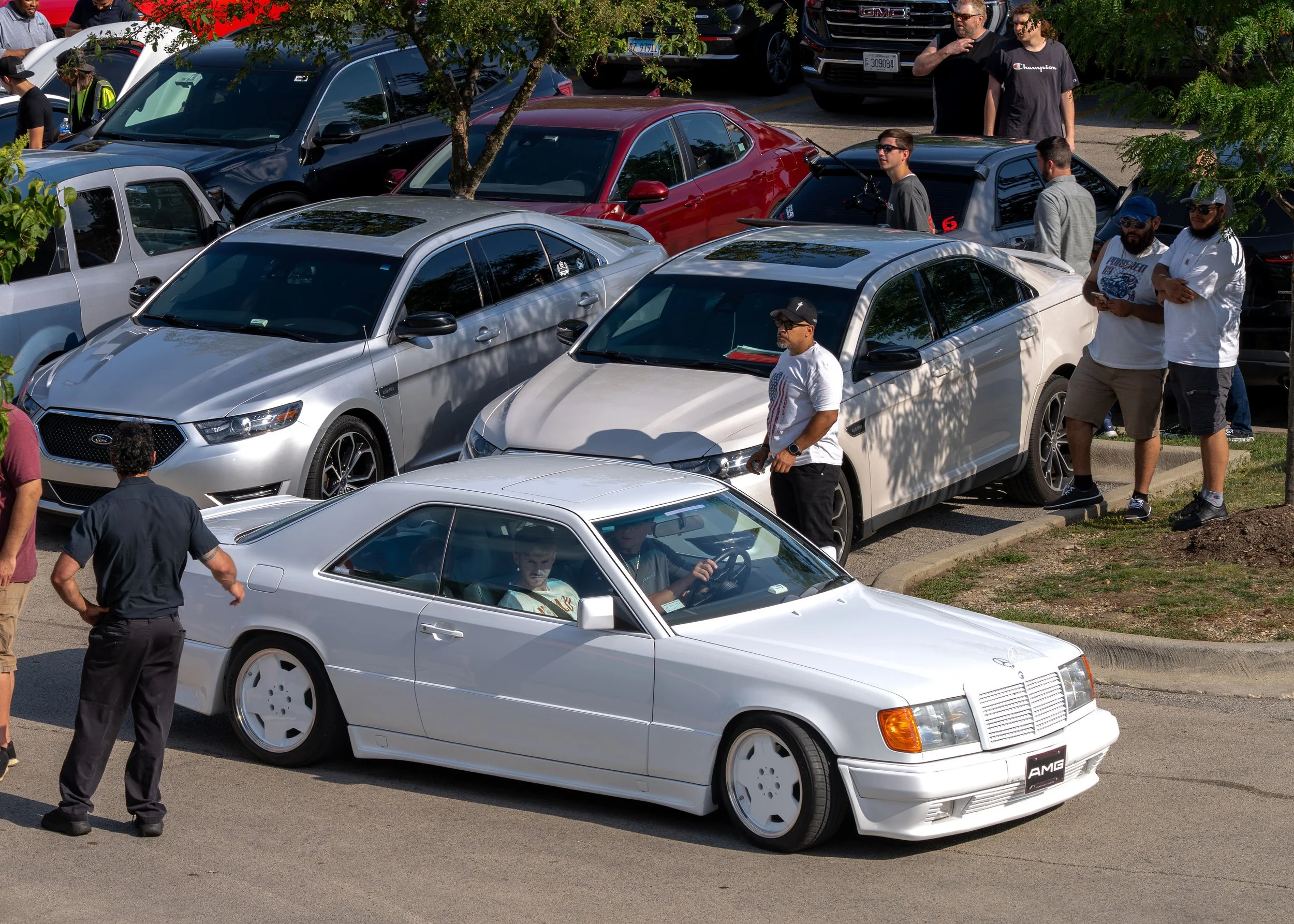 Several people gathered around parked cars in an outdoor lot, with a white sports car in the foreground and a red car behind it. Some individuals are standing near the cars, engaged in conversation, while others appear to be observing.