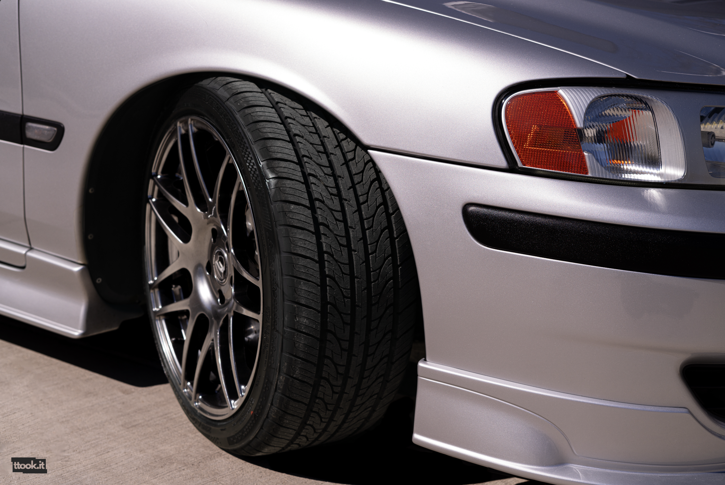 Close-up of a silver sports car's front left side, showing the wheel, tire, headlight, and part of the front bumper.