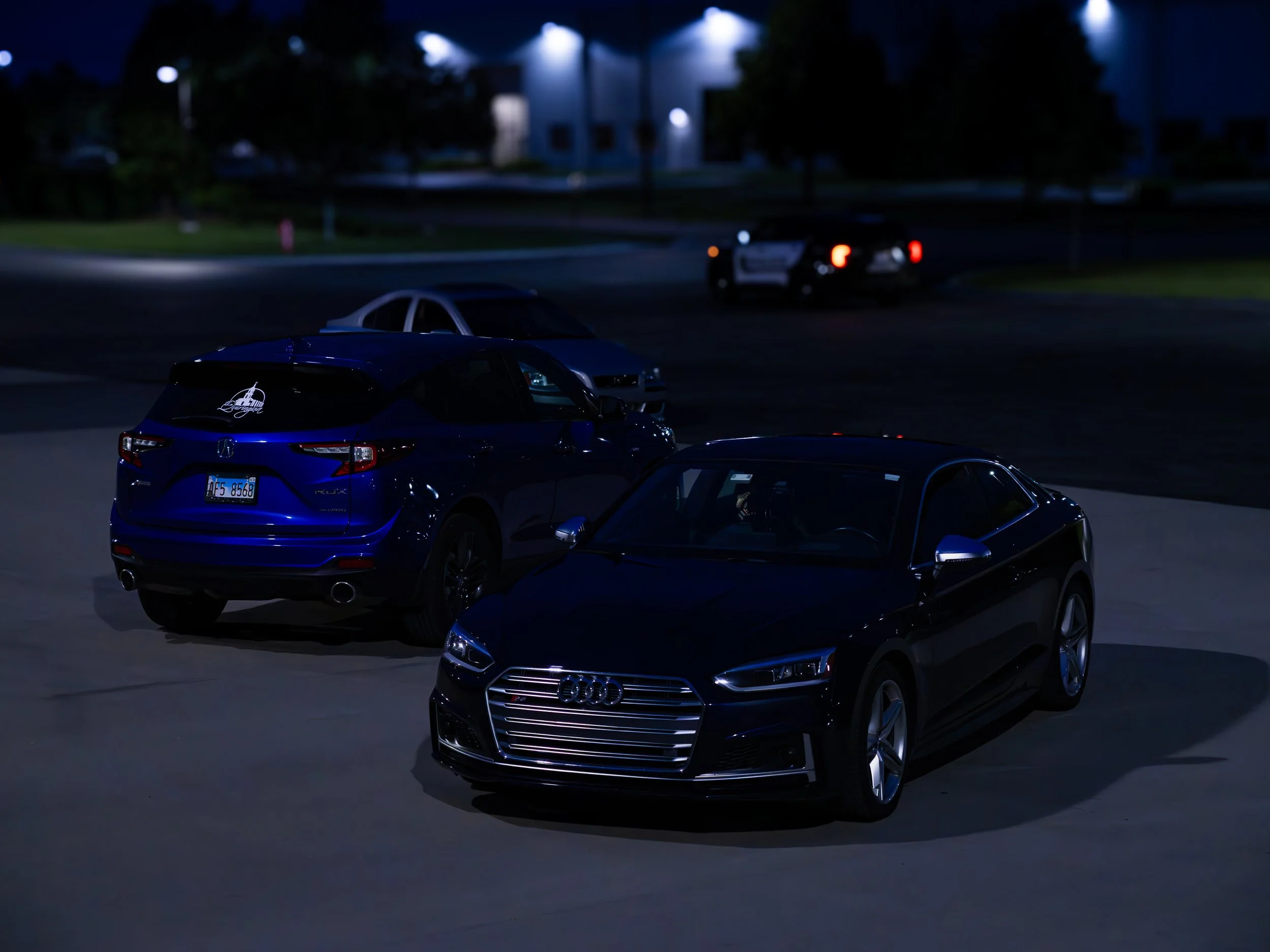 Nighttime parking lot with several parked cars, including a blue car and a black Audi. A police car with flashing lights is in the background near a building with outdoor lighting.