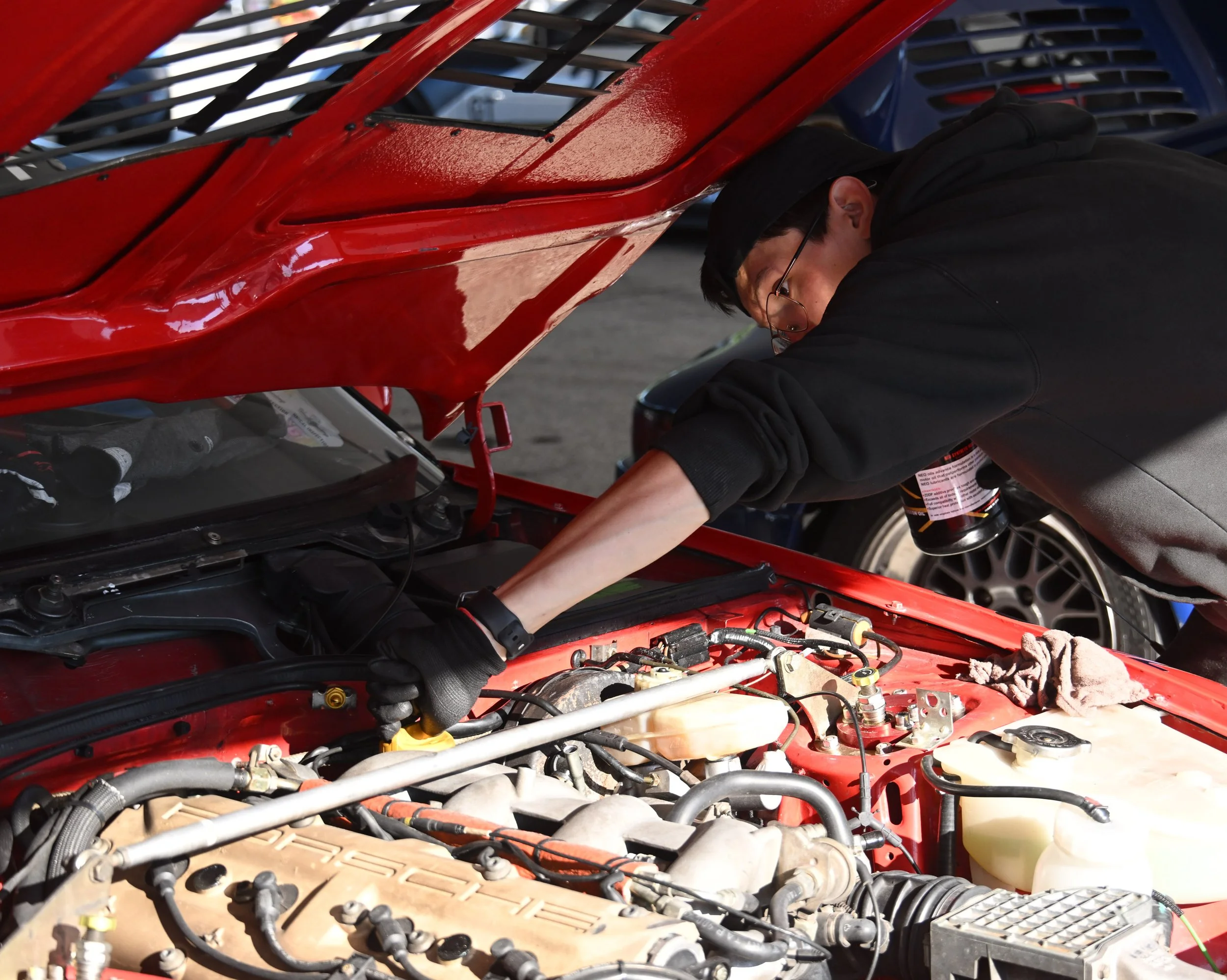 A person working on the engine of a red sports car with the hood open, glasses, black hoodie, and a cap.