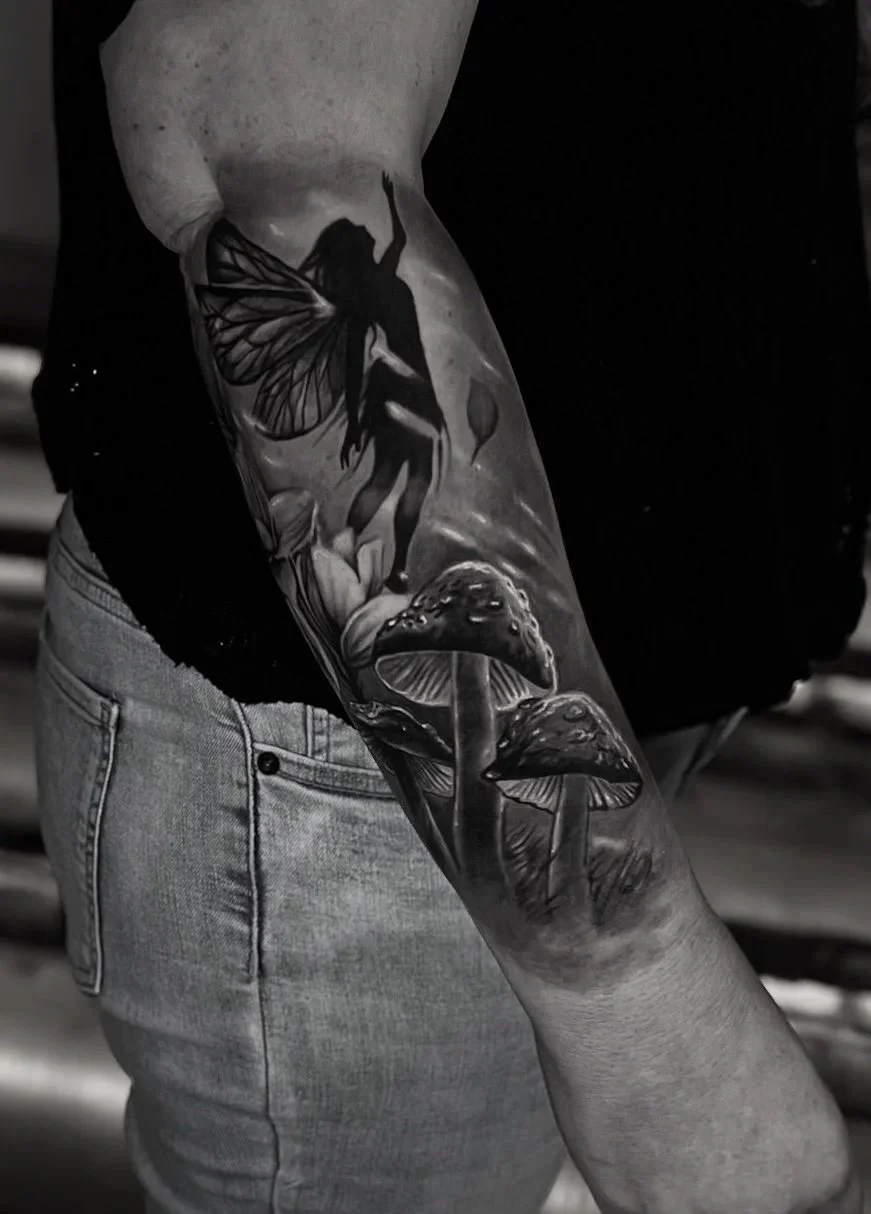 Black and white tattoo on person's arm featuring a fairy with butterfly wings, a mushroom, and botanical elements.