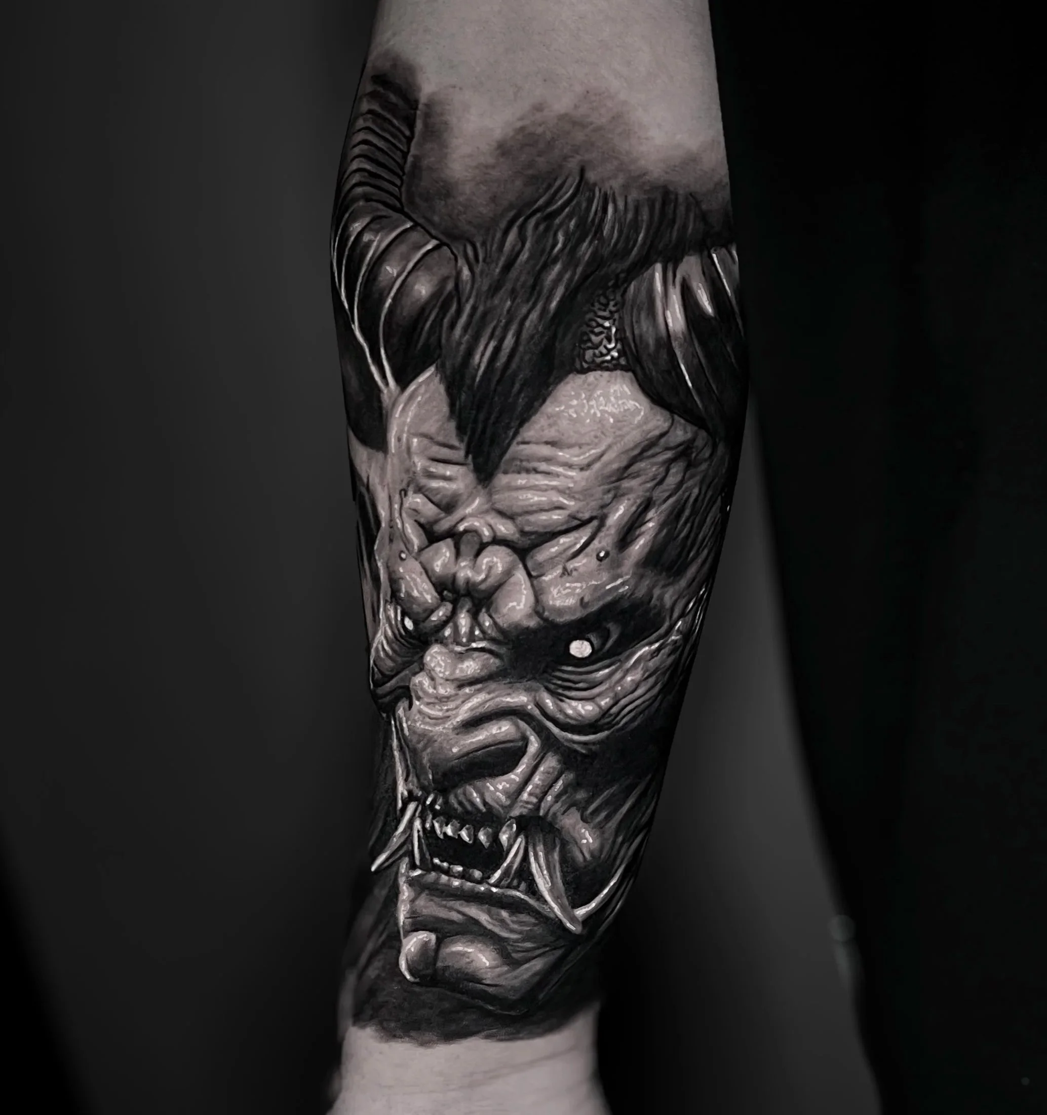 Close-up of a tattooed person's forearm featuring detailed shading and line work of a demonic face, with a dark background.