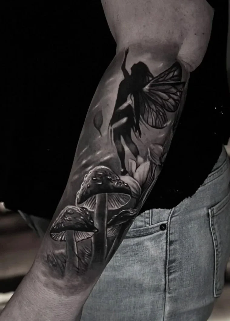 Tattoo of a fairy with butterfly wings, mushrooms, and a flower on an arm.