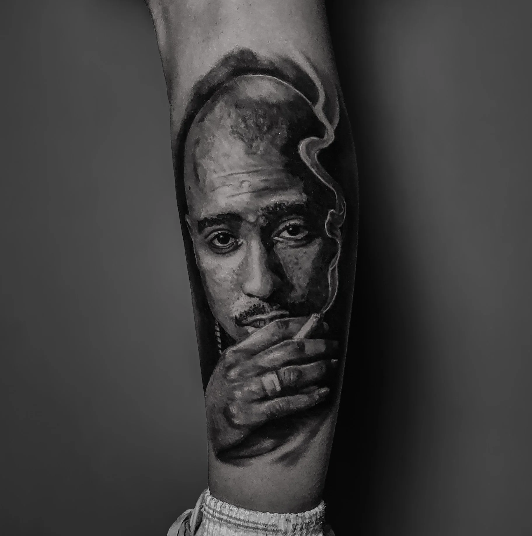 A black and white portrait tattoo Tupac on someone's arm, showing him smoking a cigarette, with detailed shading and realistic features.