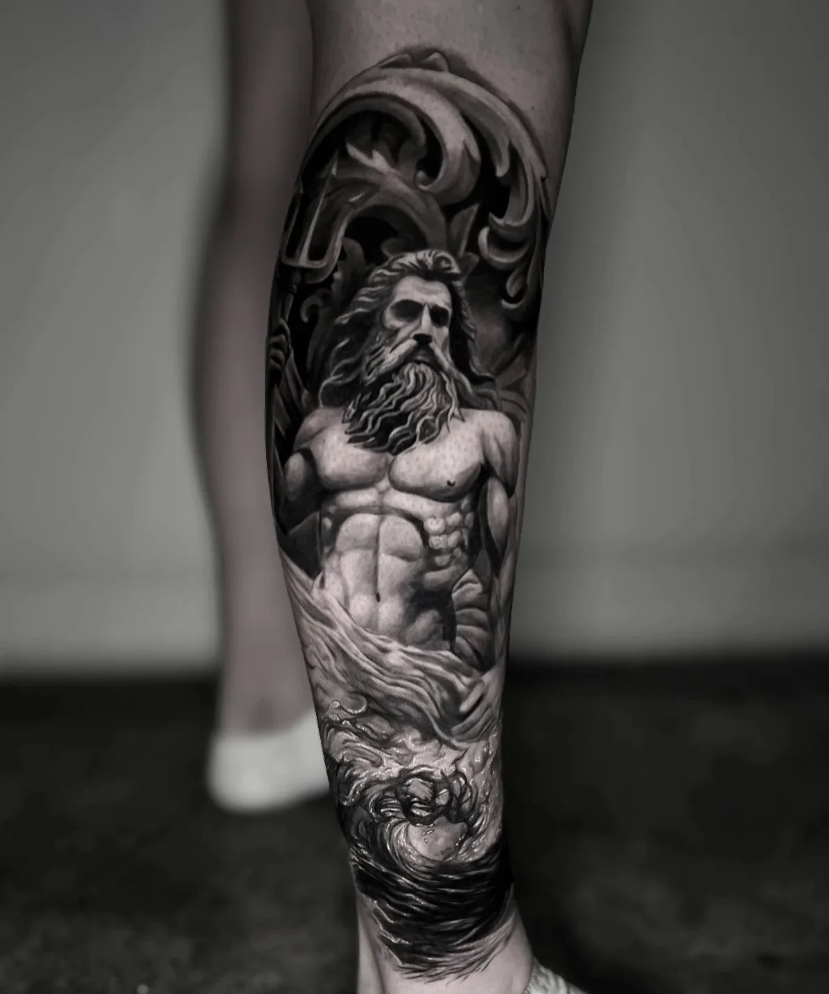 Black and gray tattoo on person's leg depicting a muscular, Poseidon with long hair, surrounded by waves and swirling filigree