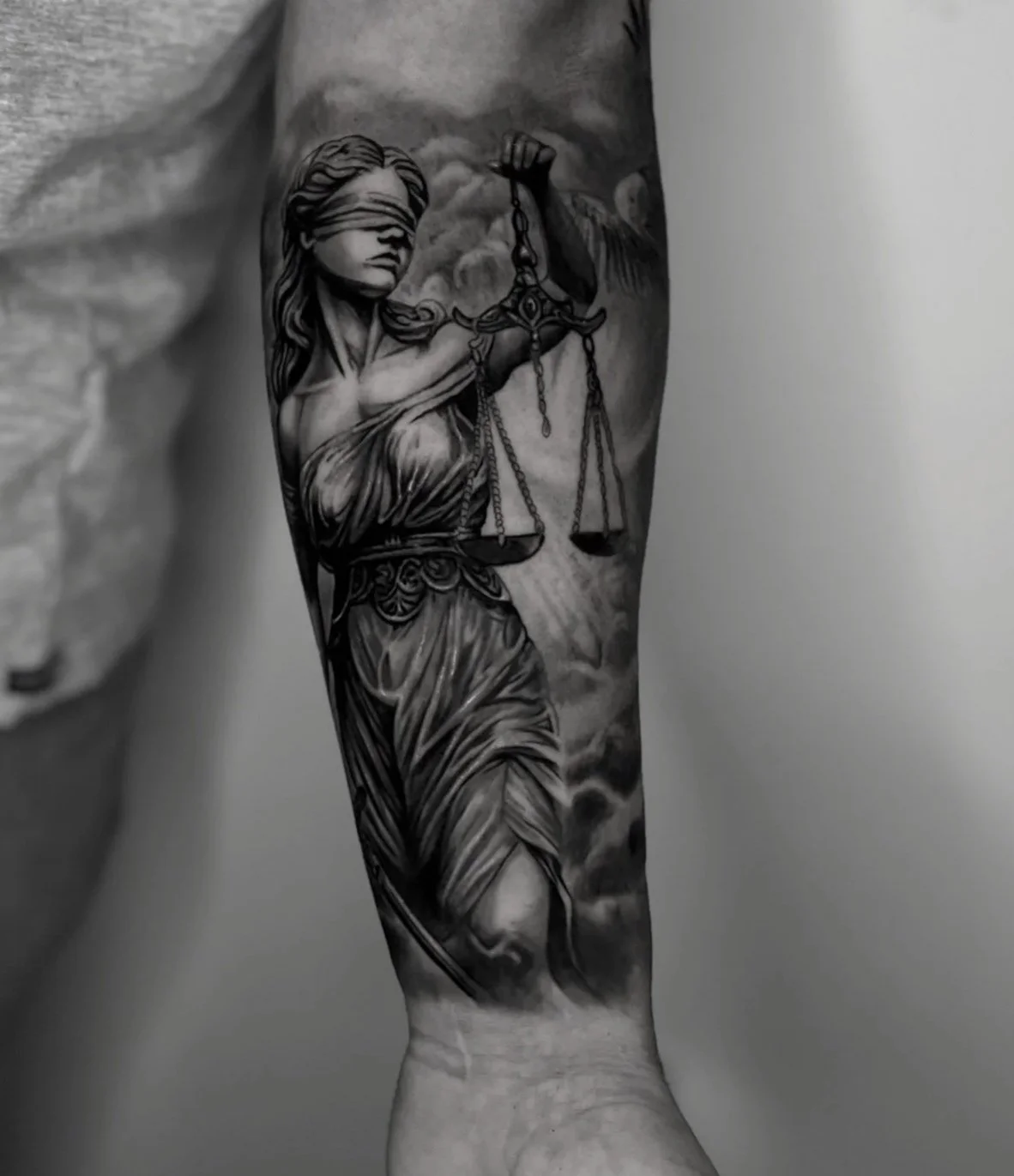 Black and gray tattoo of Lady Justice with a blindfold, holding scales in one hand and a sword in the other, on a person's forearm.