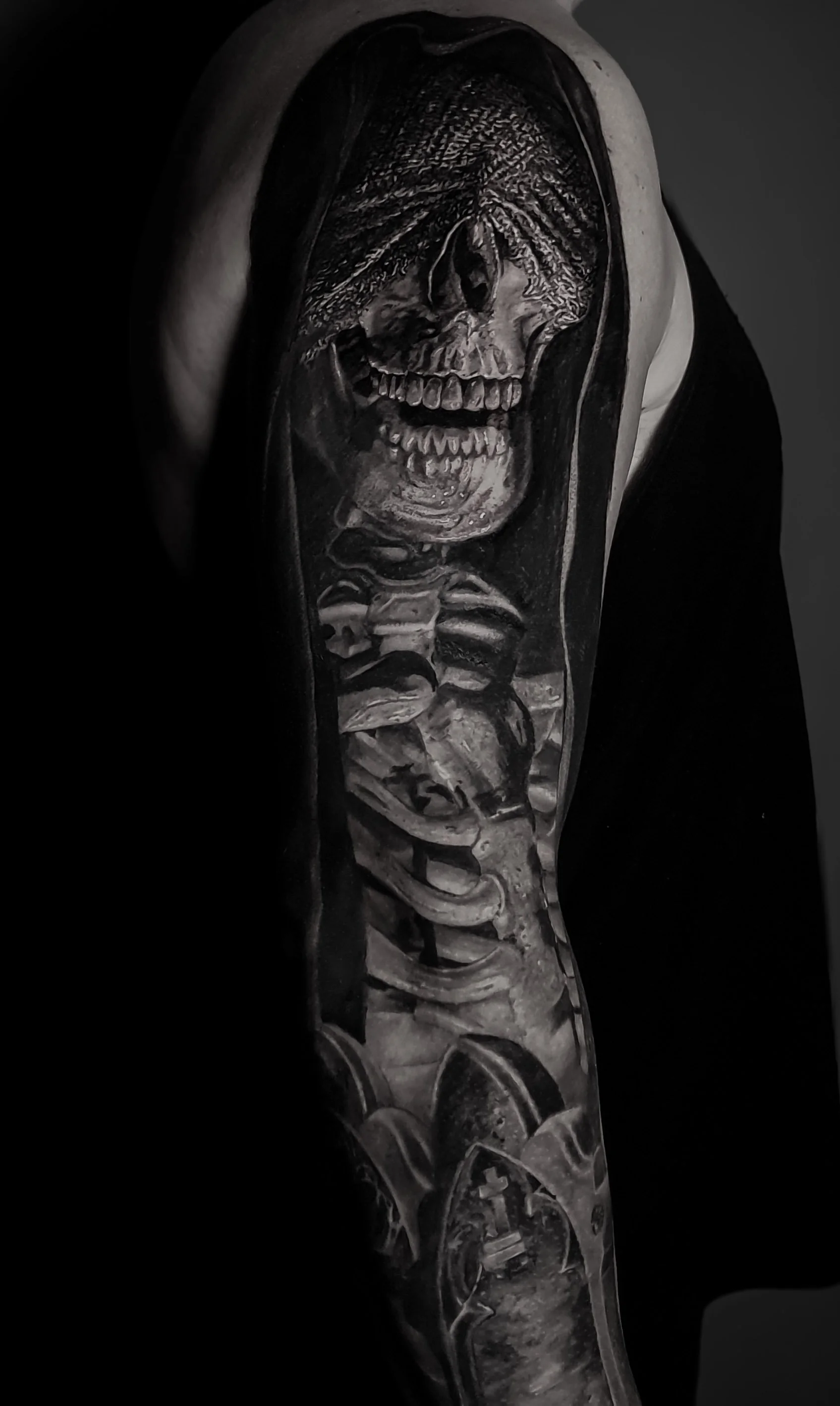 Tattoo of a skull with a hooded cloak, revealing a skeleton with visible ribs and spinal column, in black and gray ink.