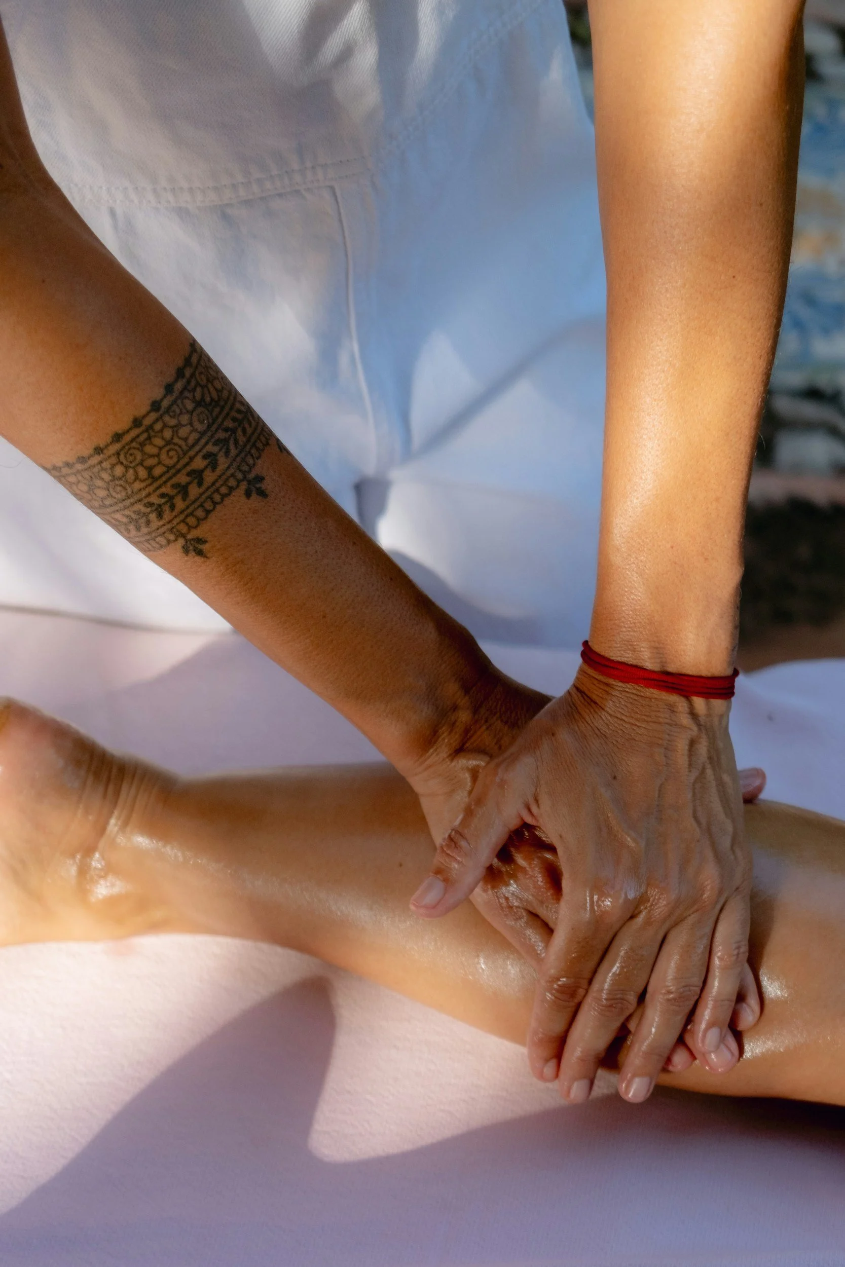 A person is receiving a massage on their leg outdoors, with the massage therapist's hands applying pressure, wearing a red bracelet and a tattoo on one arm.