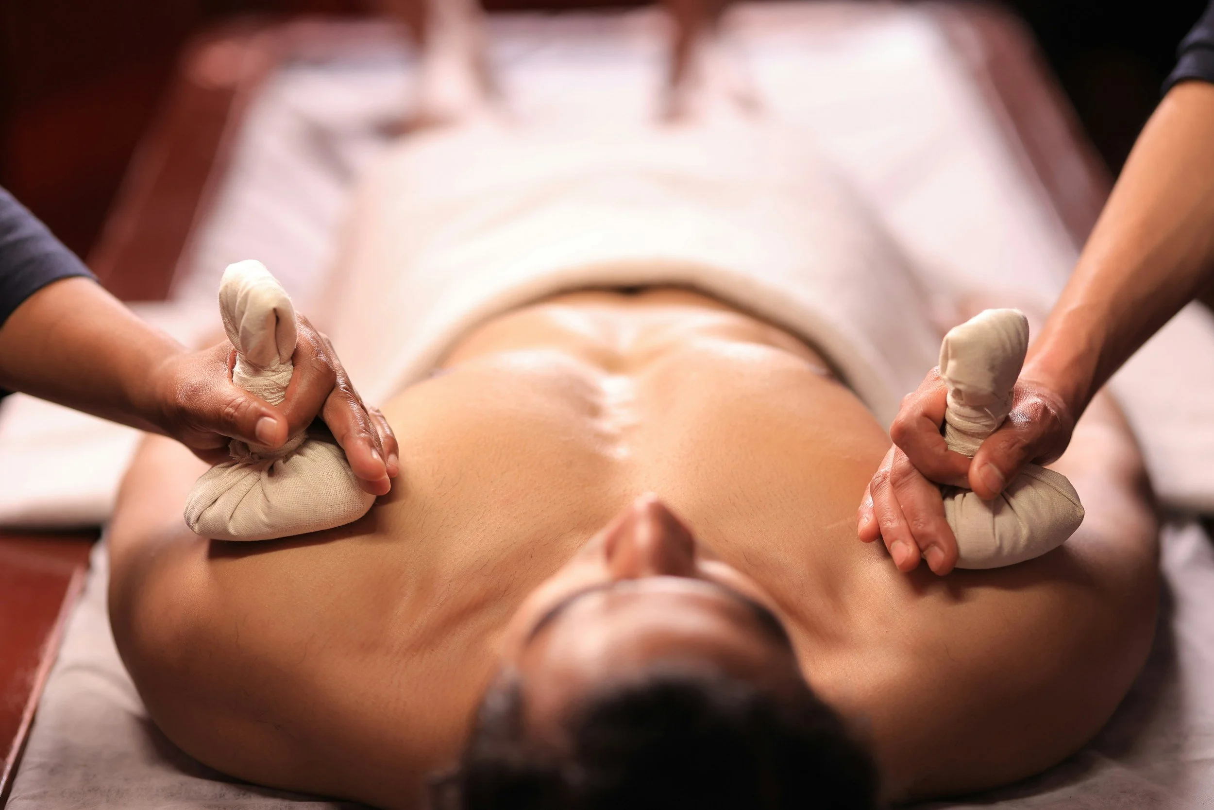 Person receiving a massage on their back with two massage therapists working on each side using herbal compresses.