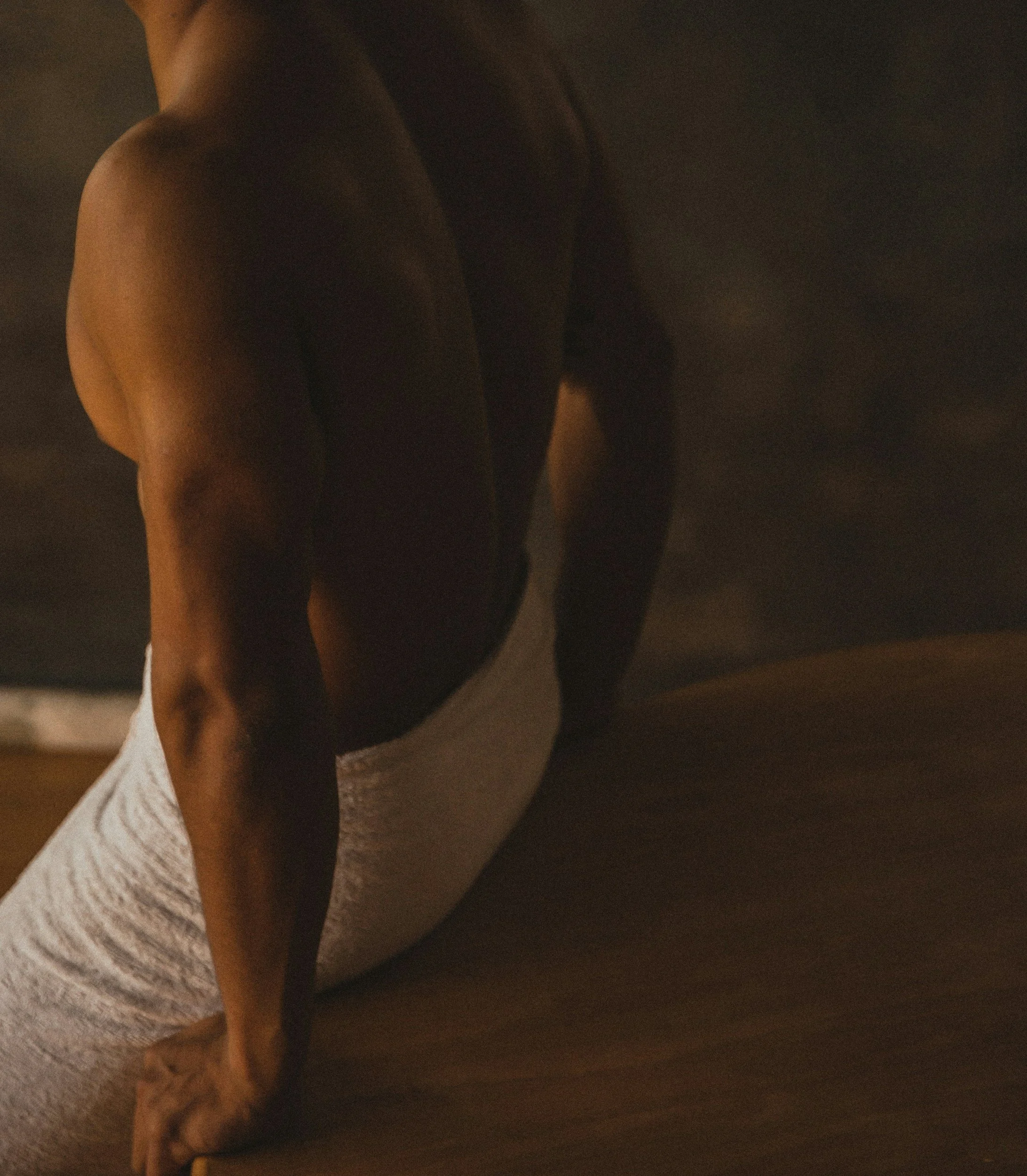A person performing a yoga pose on a wooden floor, with their body bent forward and supported by their hands and legs, in a dimly lit room after Therapeutic Massage.